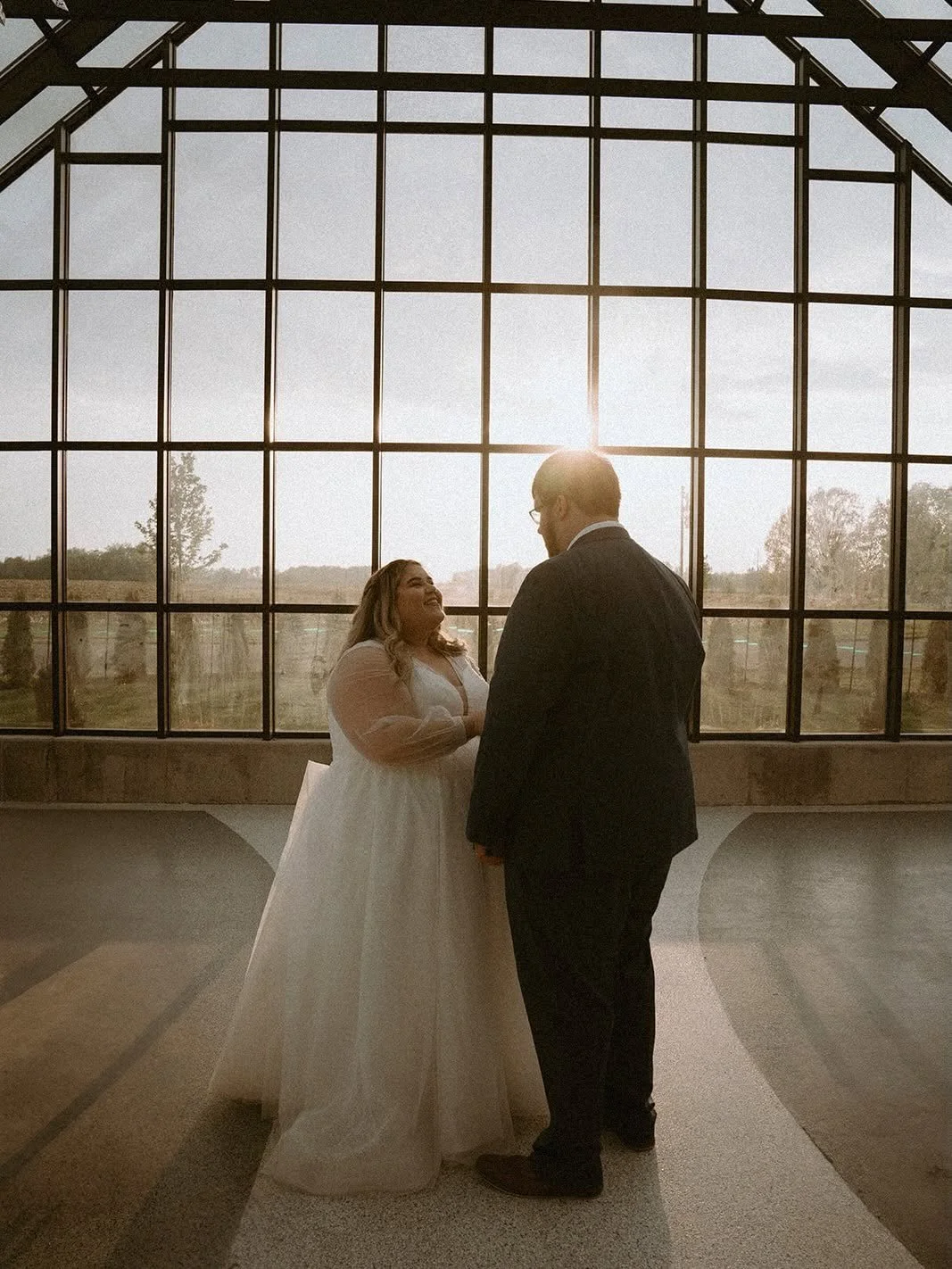 A bride and groom standing inside a building with large glass windows, holding hands and smiling at each other during sunset.