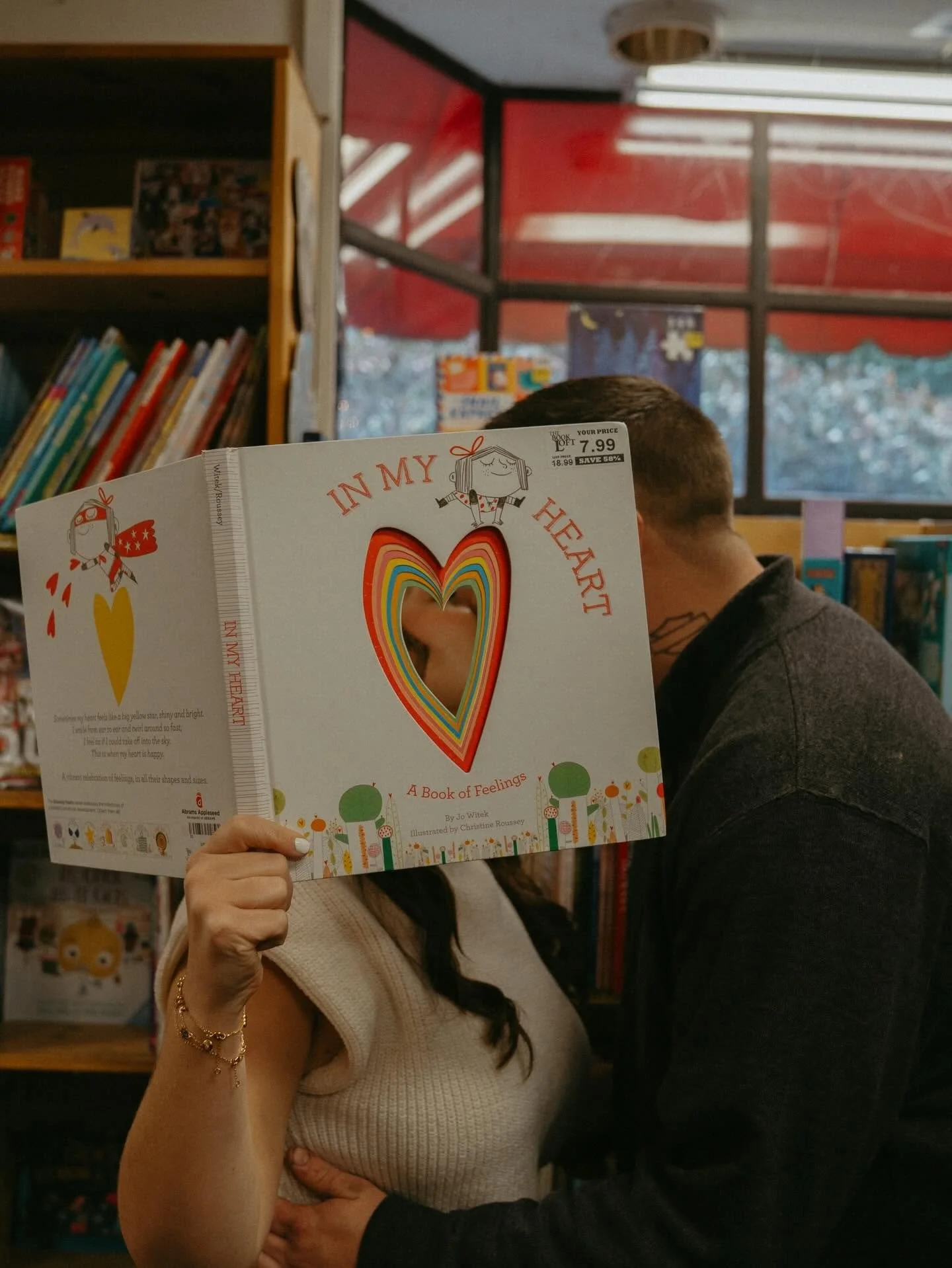 Person holding a book titled 'In My Heart' inside a bookstore, with another person standing close and partially visible. Bookshelves and an outdoor window with a red awning are in the background.