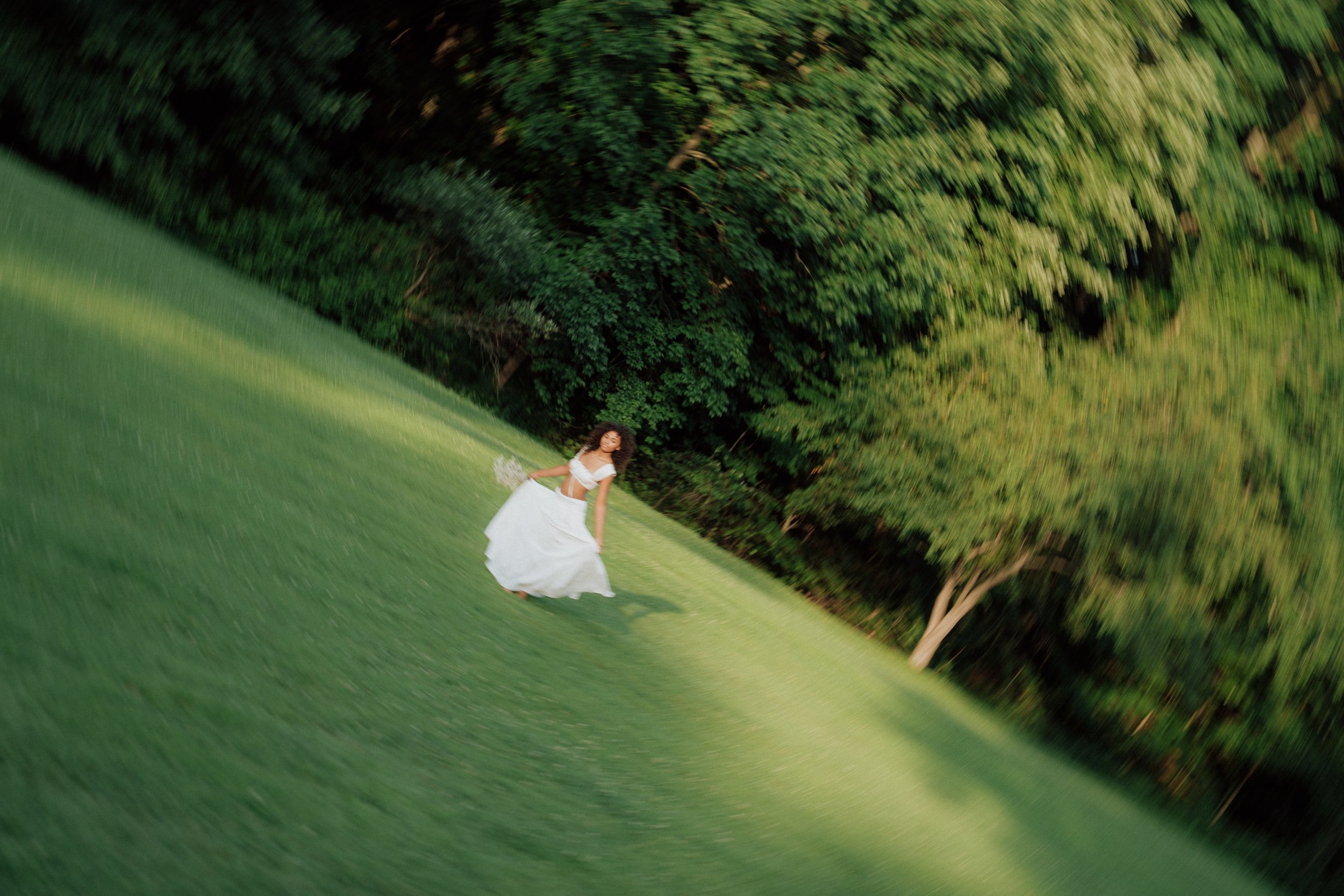 A woman in a white dress standing on a grassy field near trees, with a blurred motion effect creating a swirling pattern around her.