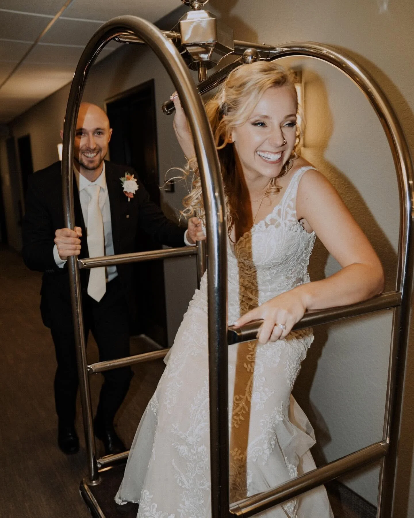 A bride with blonde hair and a big smile, wearing a white lace wedding dress, is pushing a luggage cart. A groom in a black suit and white shirt with a boutonniere is standing behind her, also smiling.