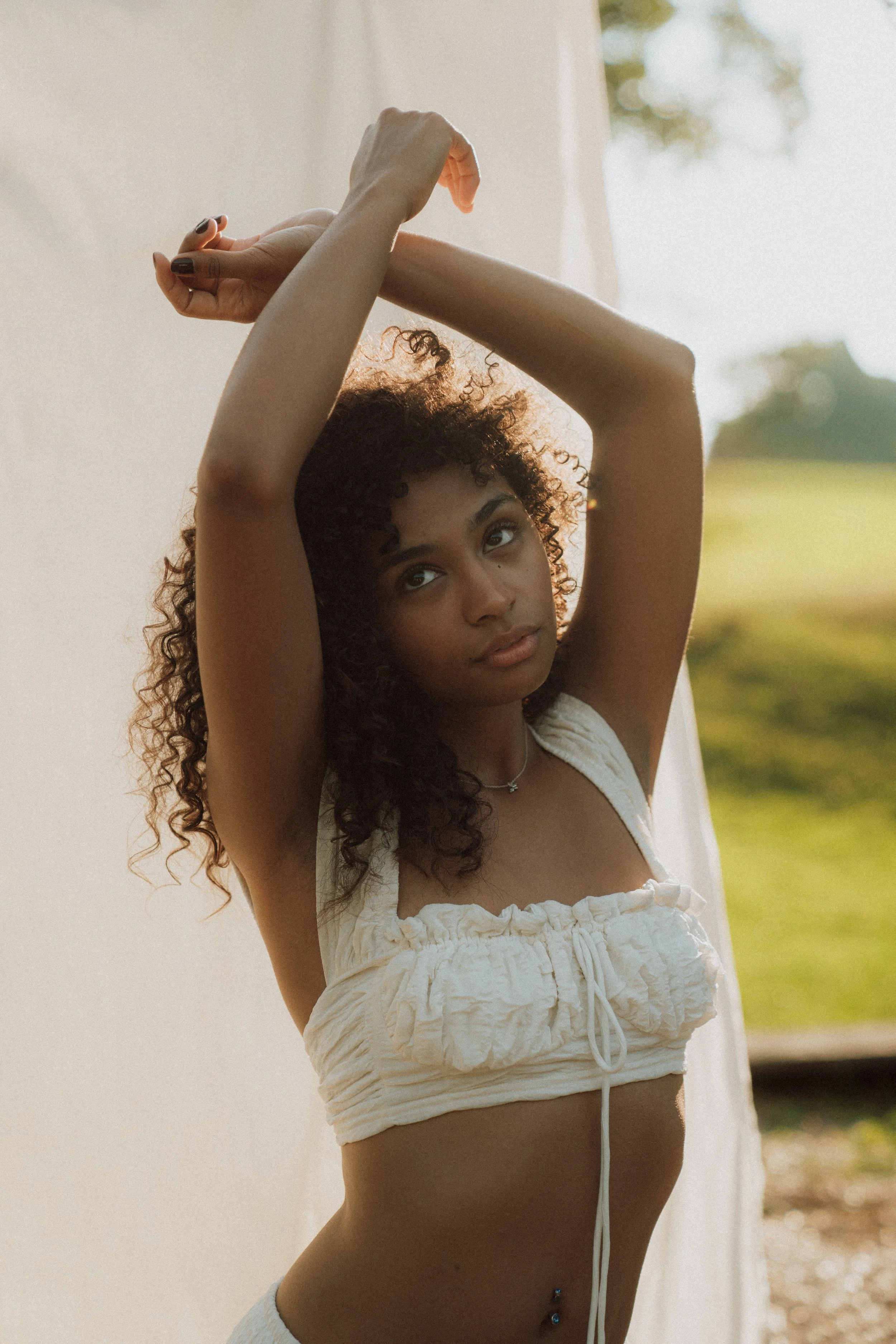 A young woman with curly hair and a belly piercing, wearing a white cropped top, has her arms raised above her head, stretching outdoors near a white wall with trees and grass in the background.