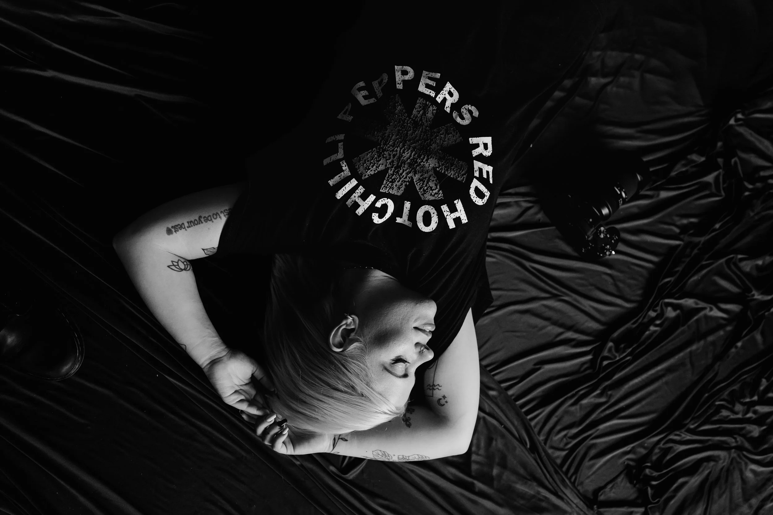 Two women lying on black fabric in a close-up shot, one with blonde hair and tattoos, smiling and looking at the other, who has dark hair, both appearing joyful and relaxed.