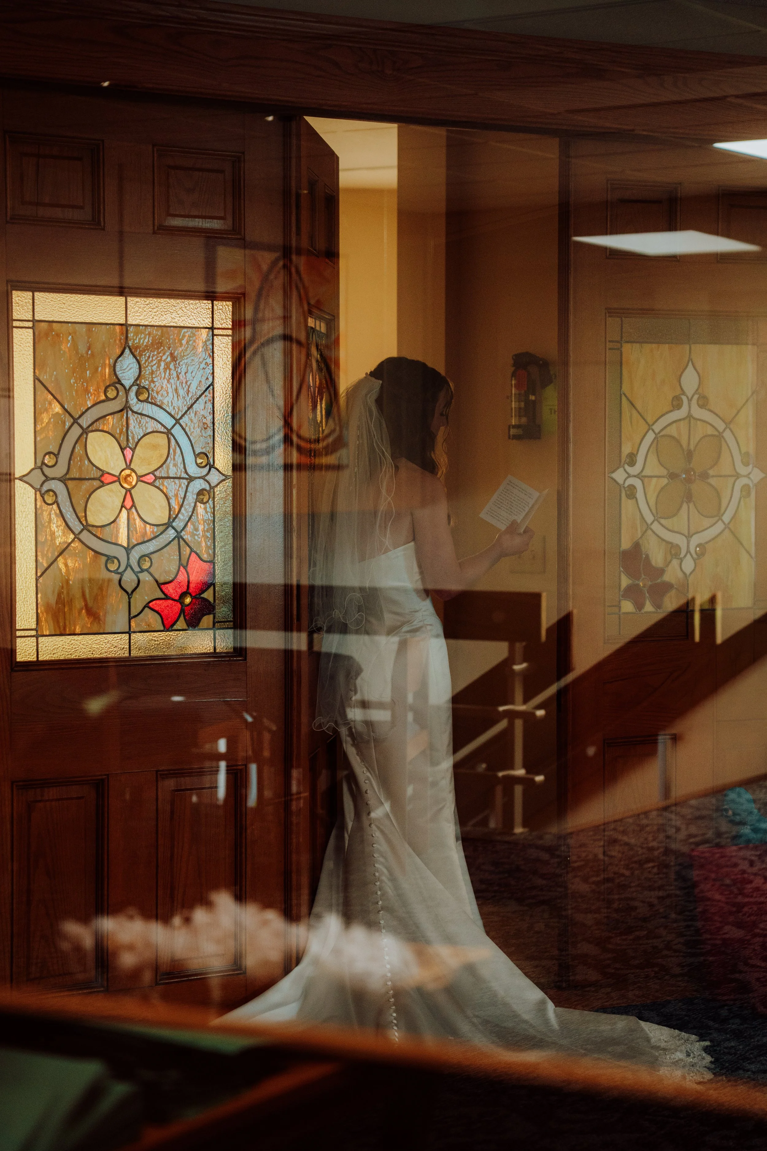 Reflection of a woman in a wedding dress holding a program, seen through a glass door with stained glass design.