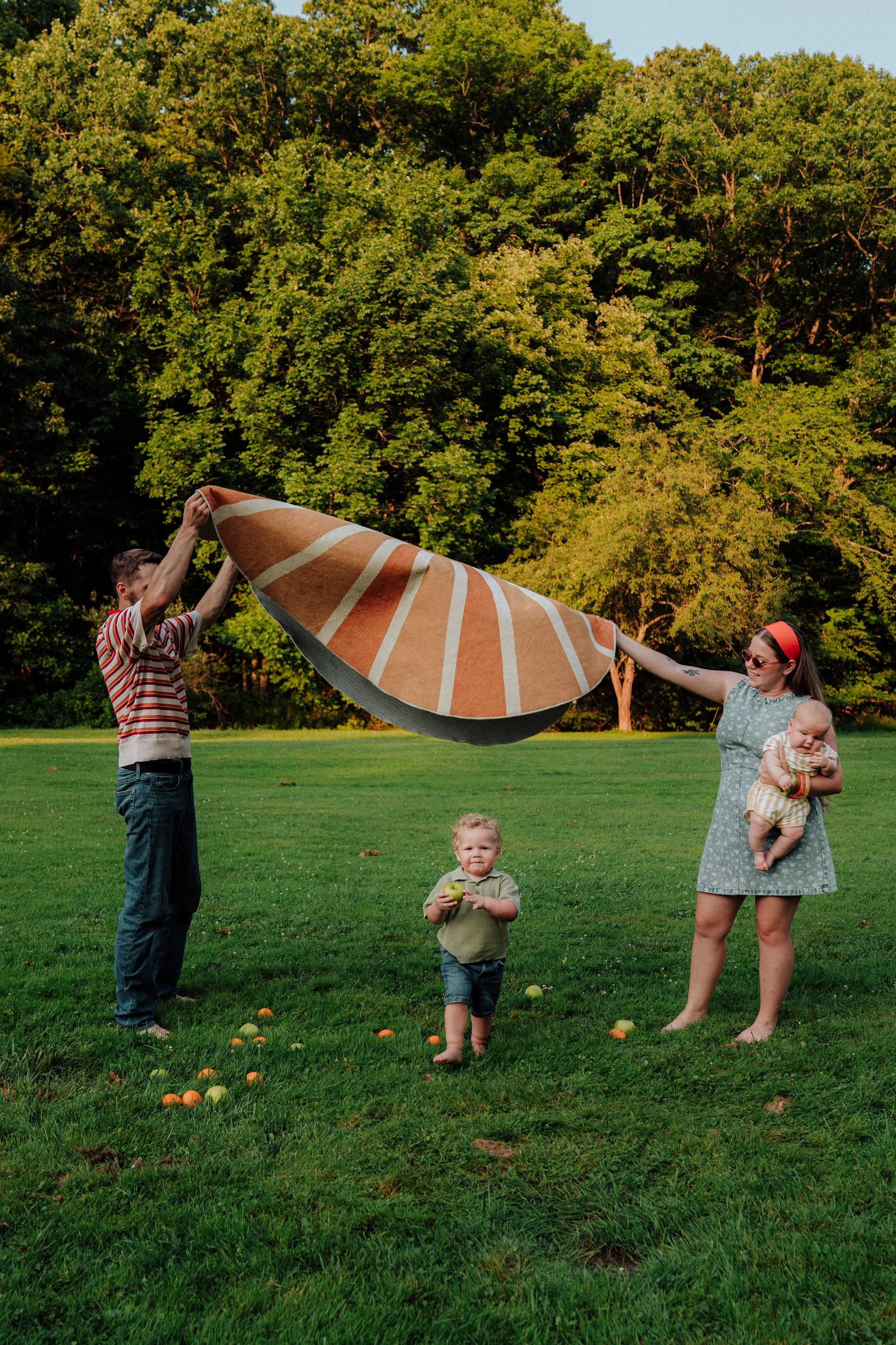 A family is outdoors in a park with green grass and large trees. A woman in a gray dress holds a baby, and a man in a striped shirt is holding a large umbrella. A young boy stands in front holding a green apple, with some oranges on the grass nearby.