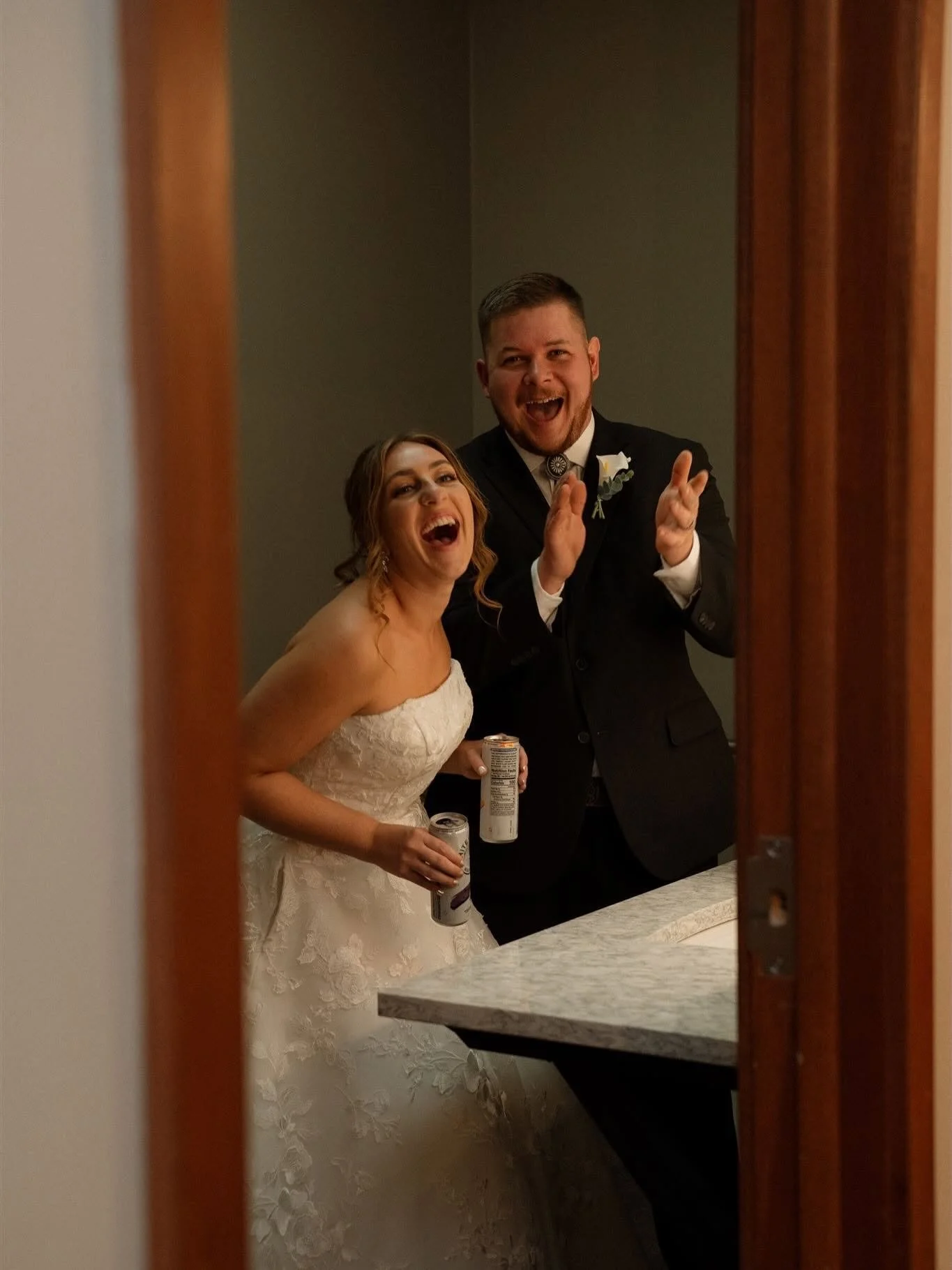 A bride and groom taking a selfie in a mirror, both smiling and holding drinks, at a wedding reception.