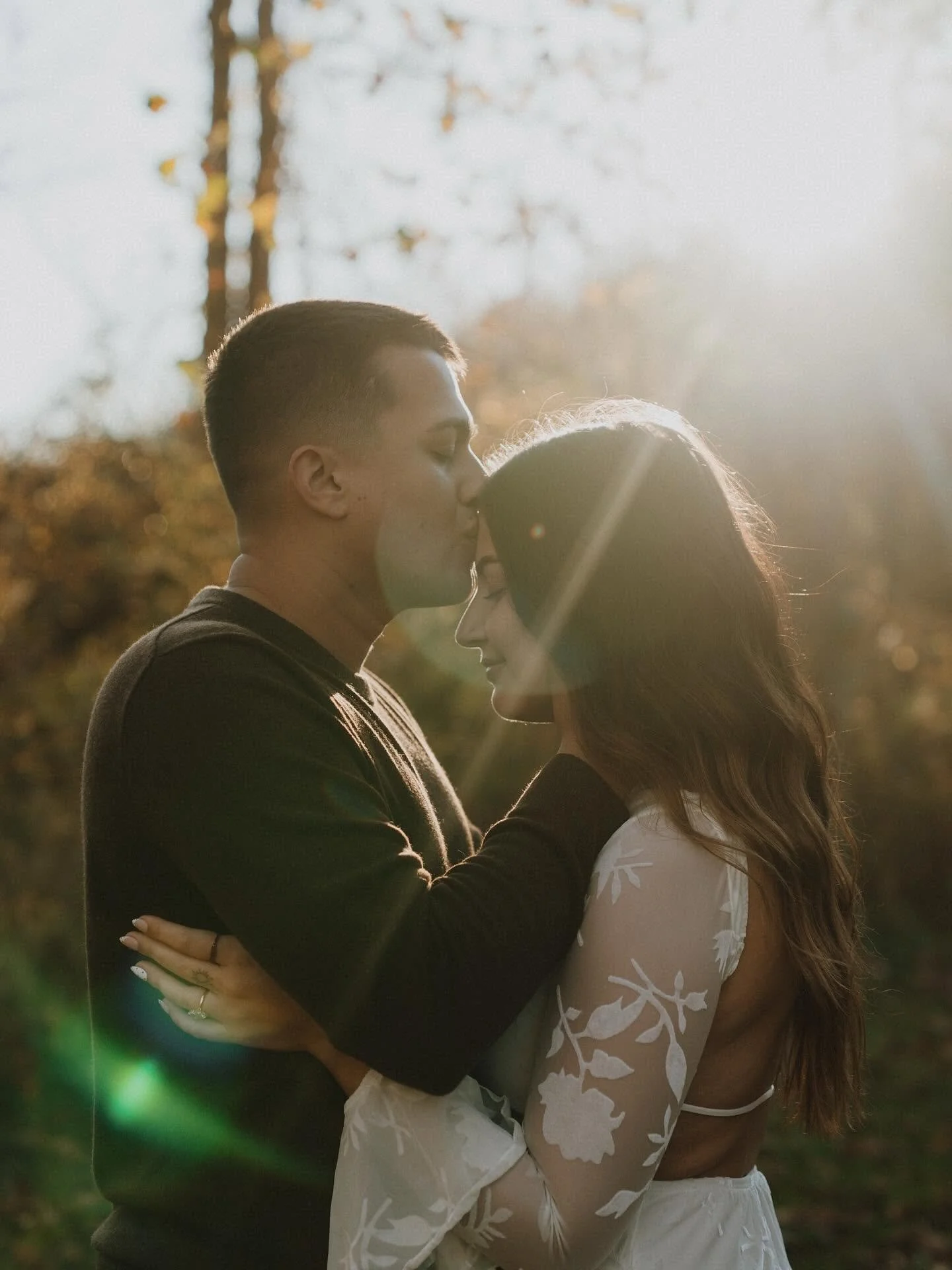 A couple is kissing outdoors in the sunlight, surrounded by trees with fall foliage.