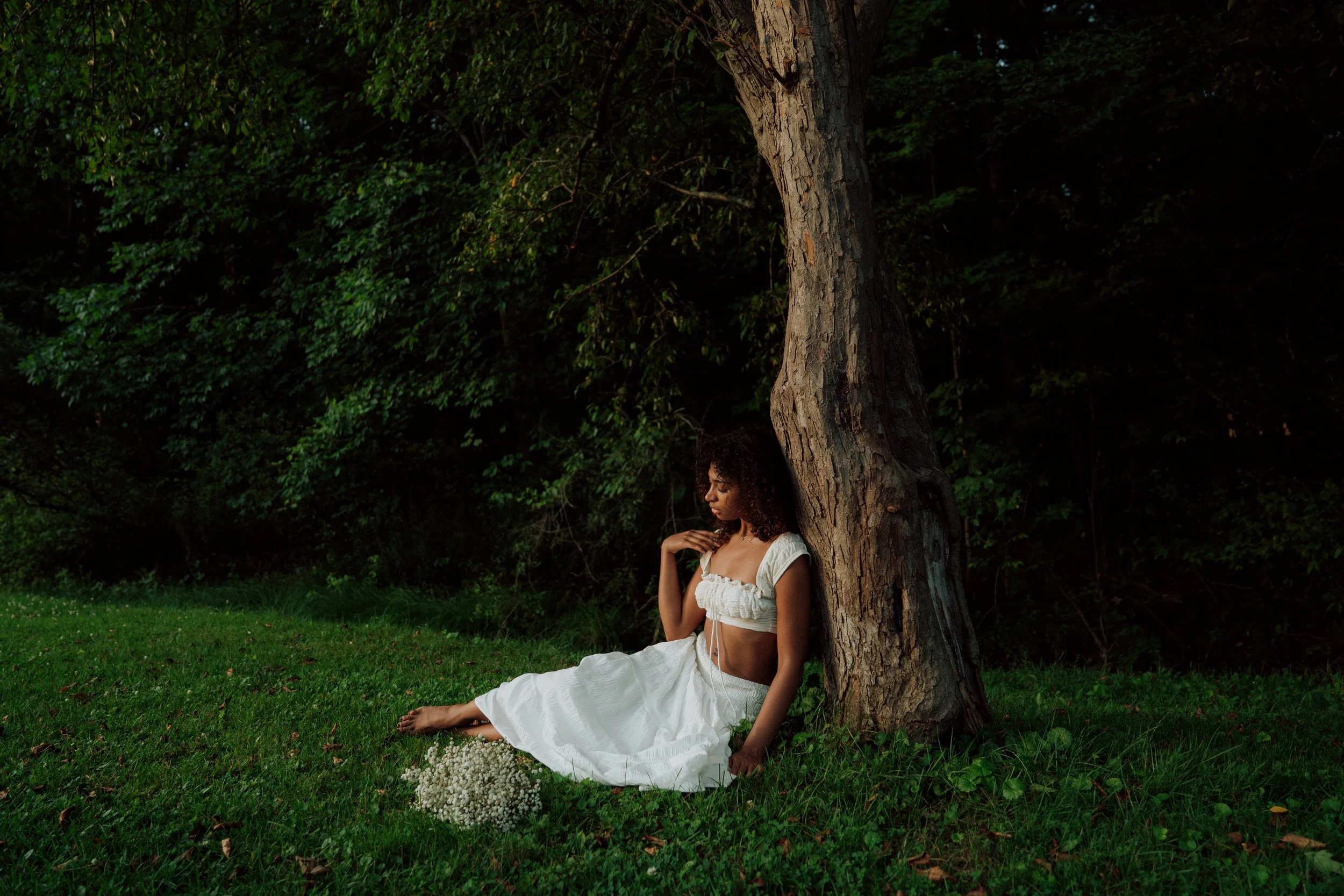 A woman with curly hair sitting under a tree in a grassy area, wearing a white dress with ruffled sleeves and a square neckline, with a bouquet of small white flowers on the ground beside her.