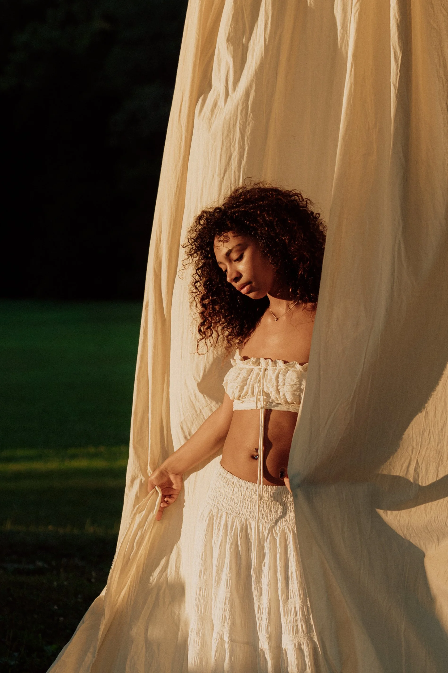 A young woman with curly hair wearing a beige, textured two-piece outfit standing in front of a large cream-colored fabric backdrop at night, with greenery and darkness in the background.