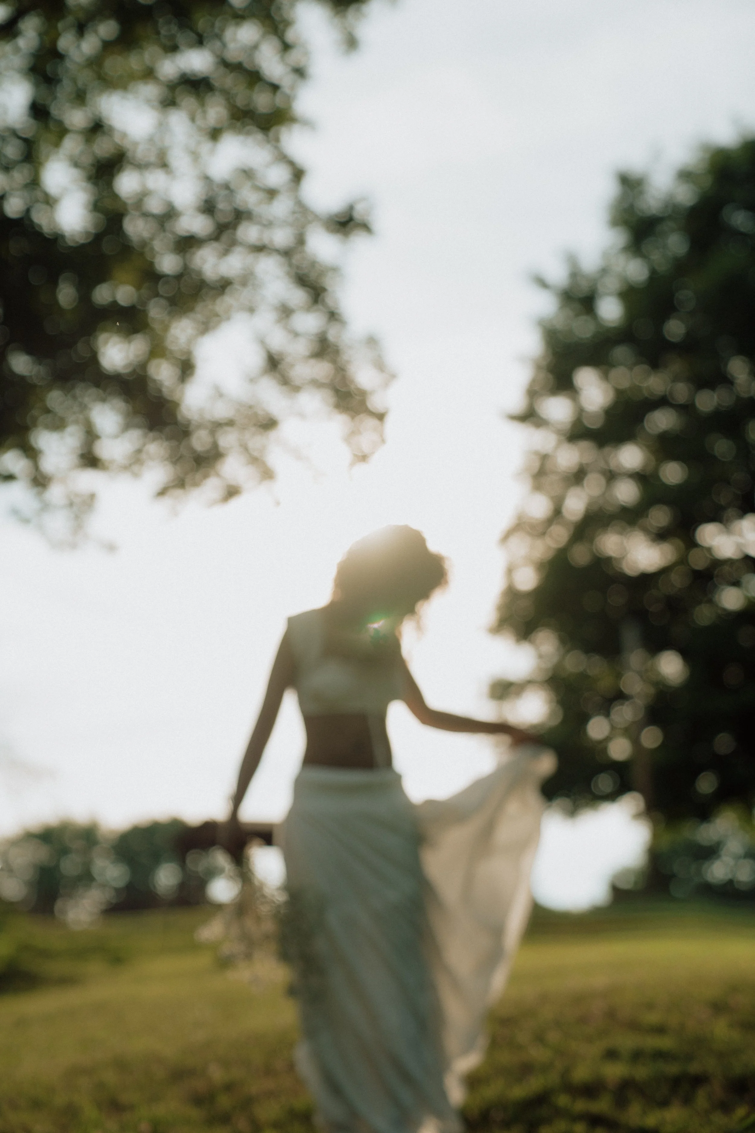 A person in light-colored clothing is dancing outdoors under trees with the sun shining through the leaves.