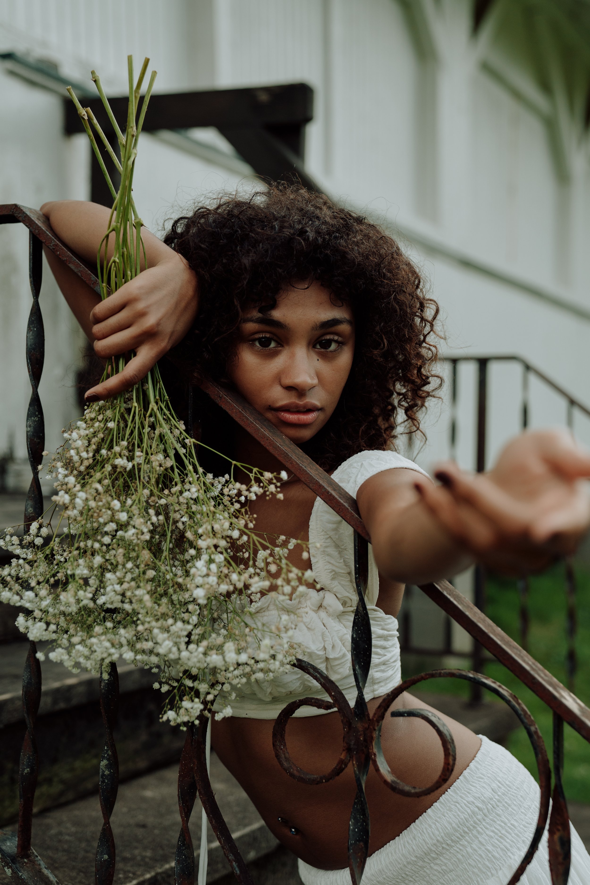 A young woman with curly hair and light brown skin, wearing a white dress, sitting on outdoor stairs behind a black wrought iron fence, holding a bouquet of small white flowers.