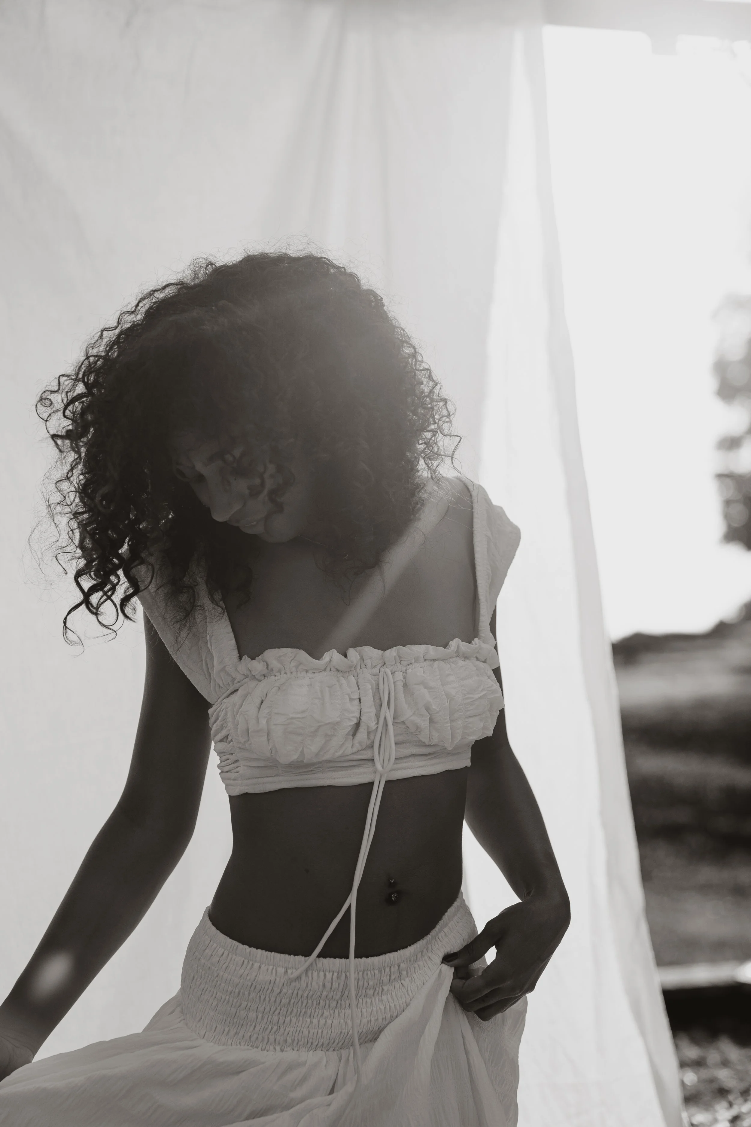 A woman with curly hair wearing a white crop top and a long skirt, standing outdoors near a white curtain or sheet, in black and white.