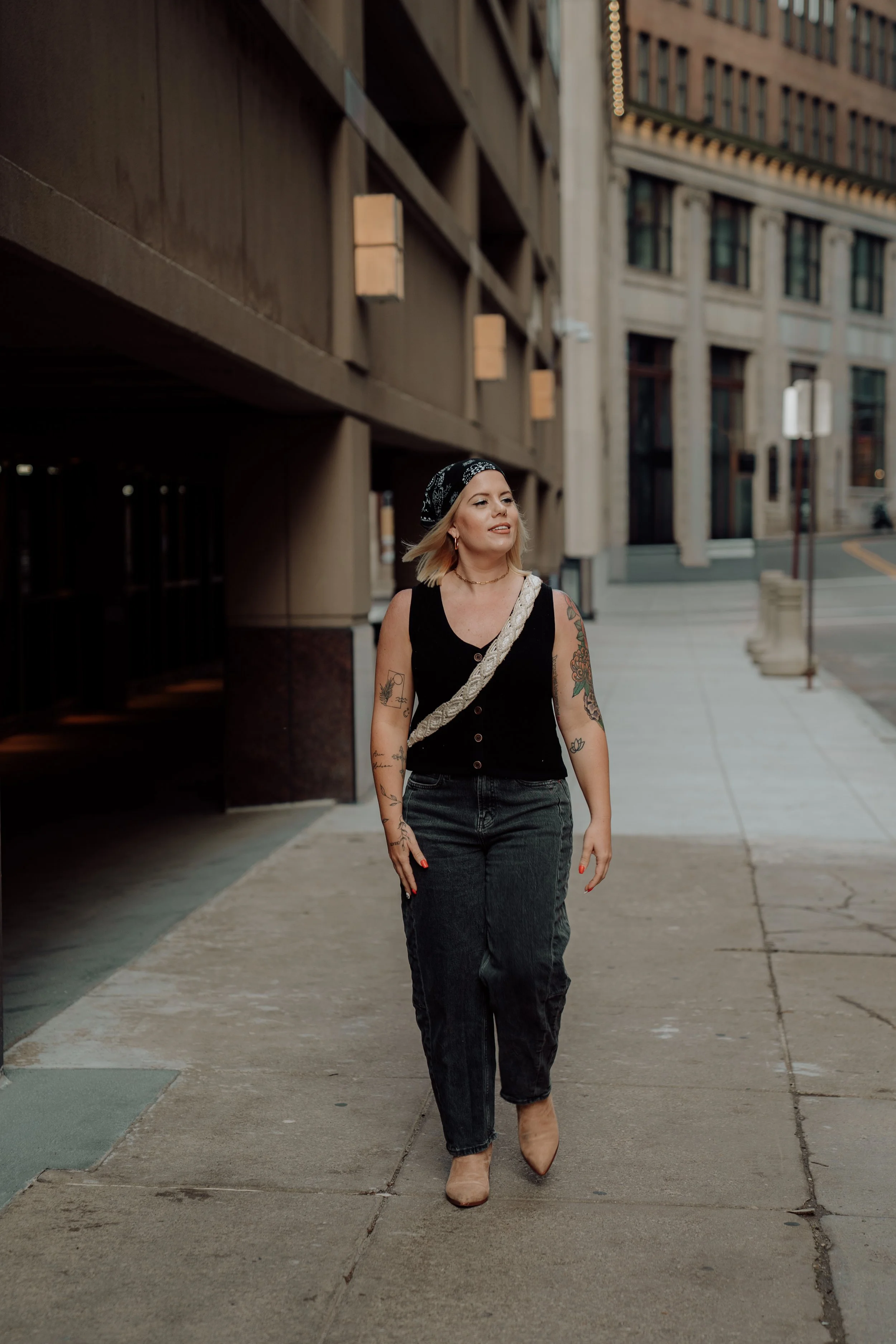 A woman with tattoos on her arms walking on a city sidewalk, wearing a black sleeveless top, dark jeans, beige shoes, a black bandana, and a beige strap bag, with tall buildings in the background.