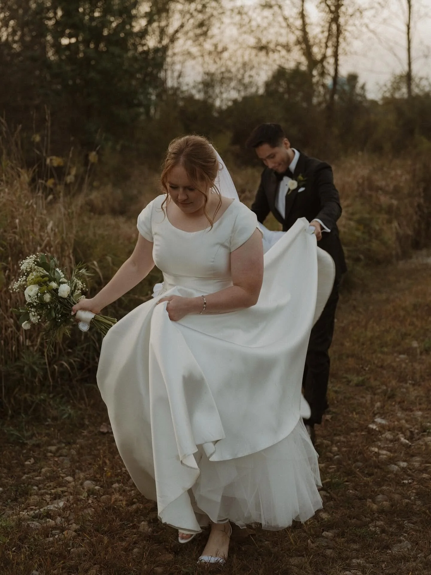 A bride in a white wedding dress holding a bouquet, with a groom in a black tuxedo behind her, in an outdoor setting with trees and grass during sunset.