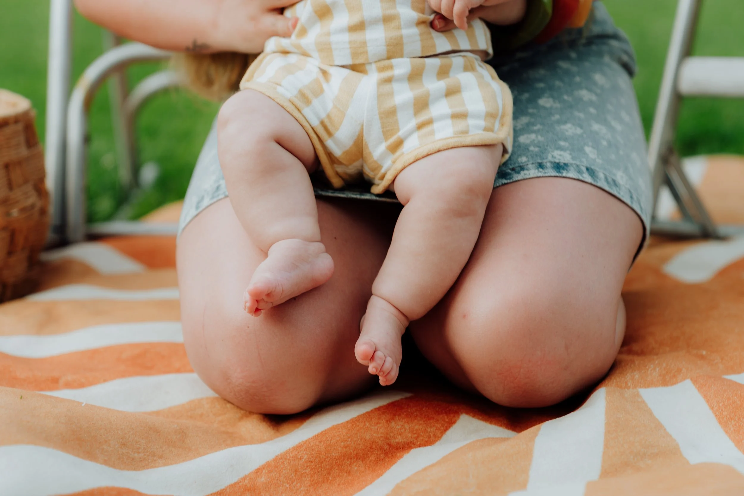 Close-up of a baby wearing a yellow and white checkered outfit, being held with adult hands, and sitting on an orange and white patterned blanket outdoors.