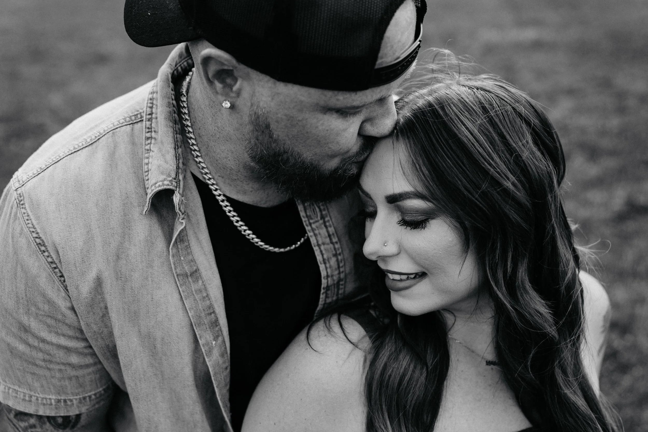 A black-and-white photograph of a man with a beard, wearing a baseball cap, earrings, a chain necklace, and a denim jacket, embracing a woman with long hair, closed eyes, and a subtle smile.