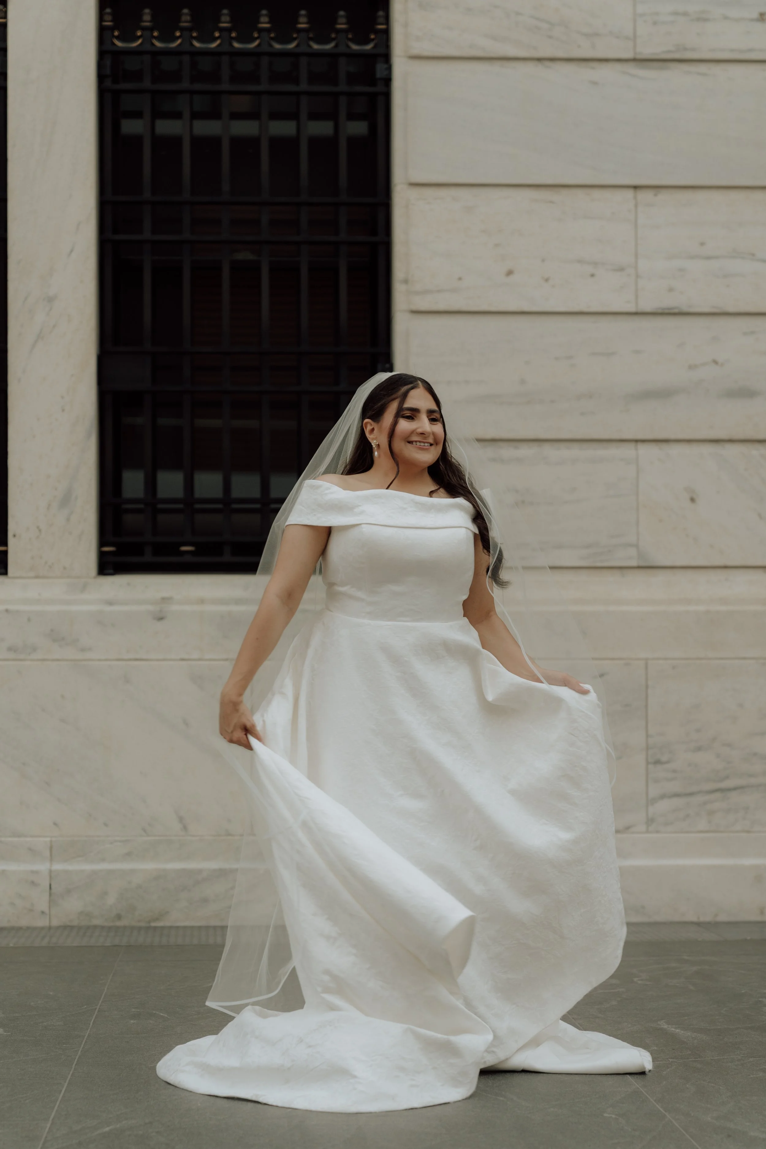 A woman in a white wedding dress holding part of her dress, standing outside in front of a marble wall with a black barred window, smiling.