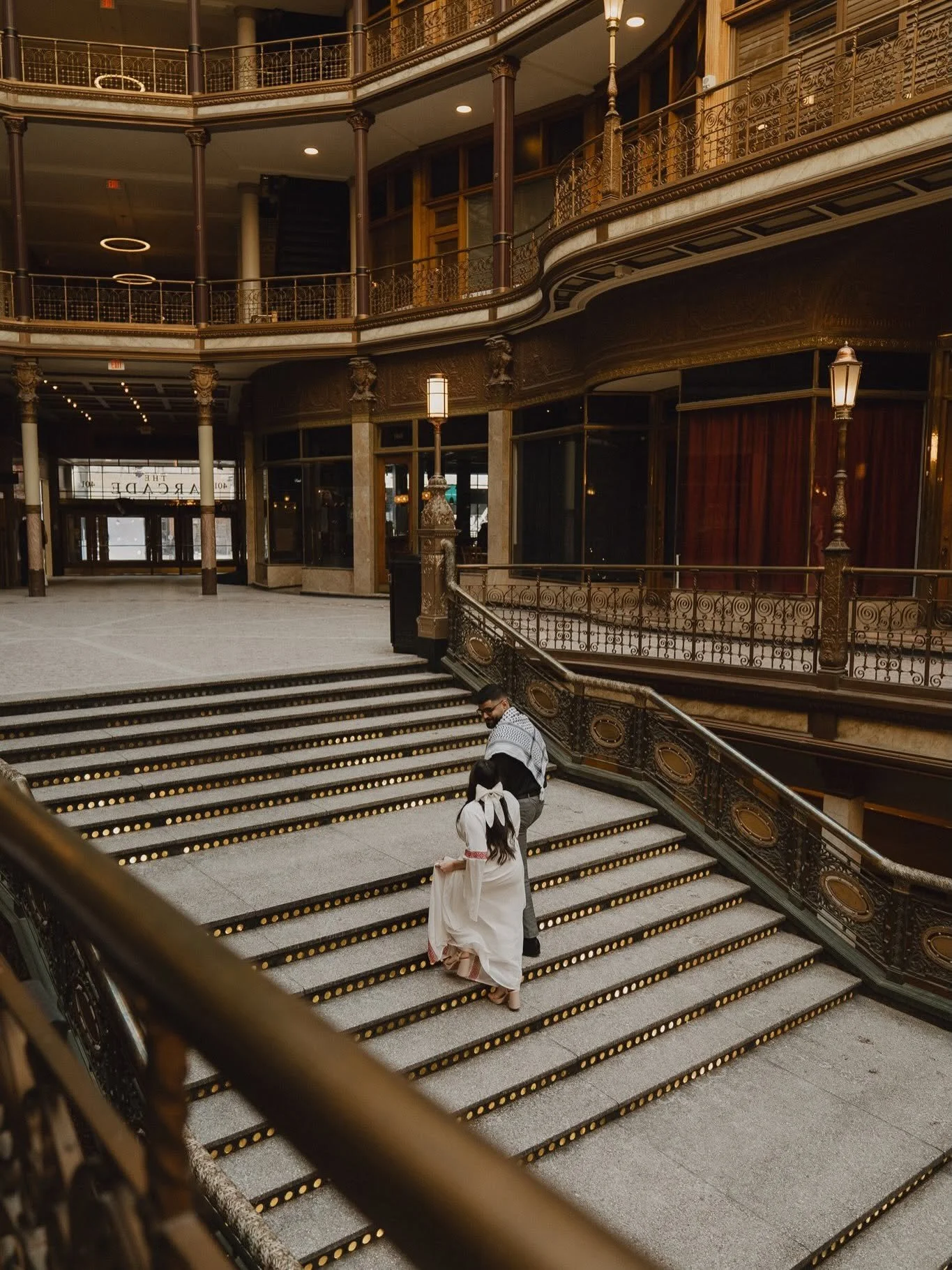 A man and woman in formal attire walking up a grand staircase inside a historic building with ornate railings, gold accents, and tall lamps, possibly during a wedding or special event.