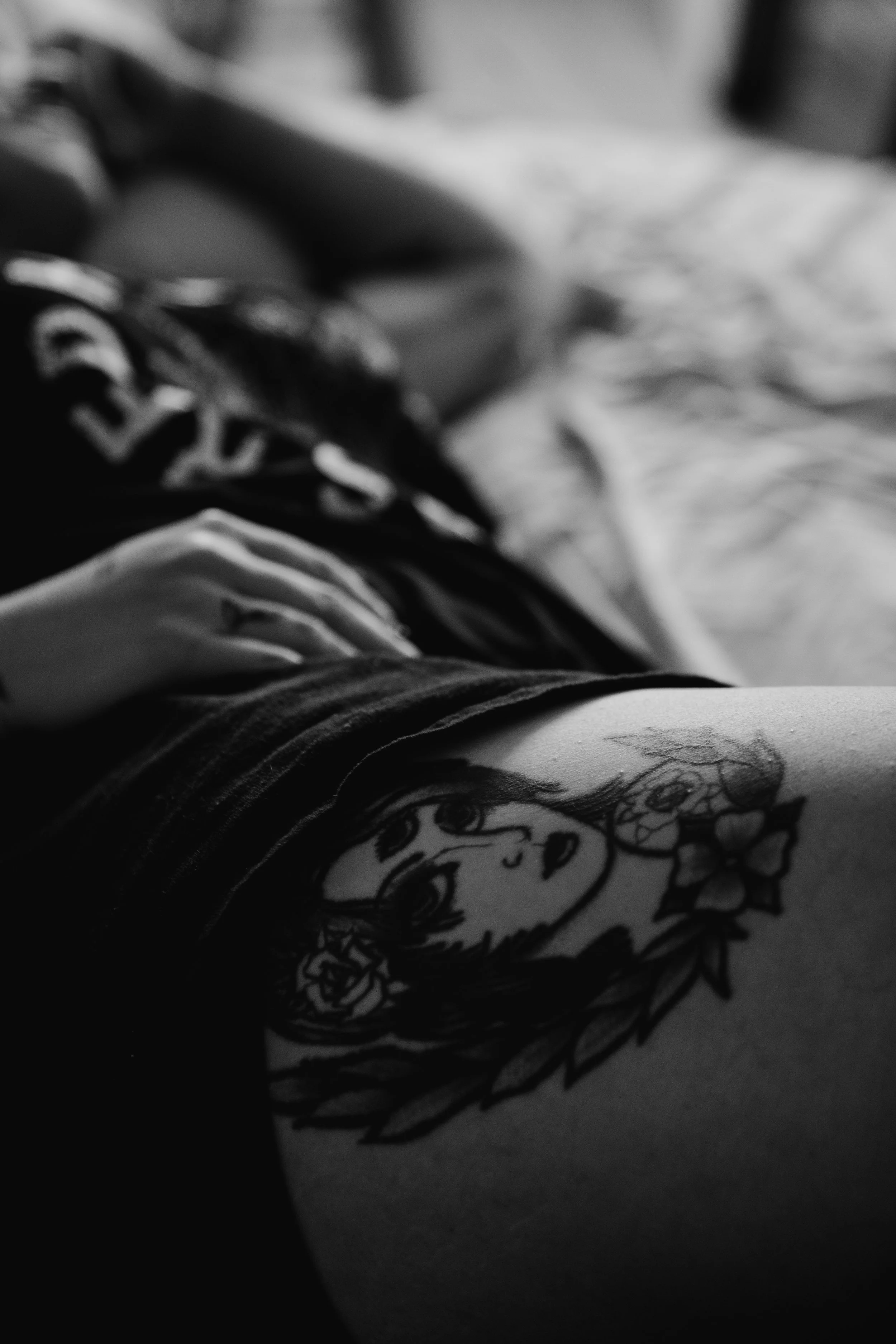 A black and white close-up photo of a tattoo on a person's skin, featuring a detailed cartoon-style face with large eyes and an expressive mouth, surrounded by floral elements.