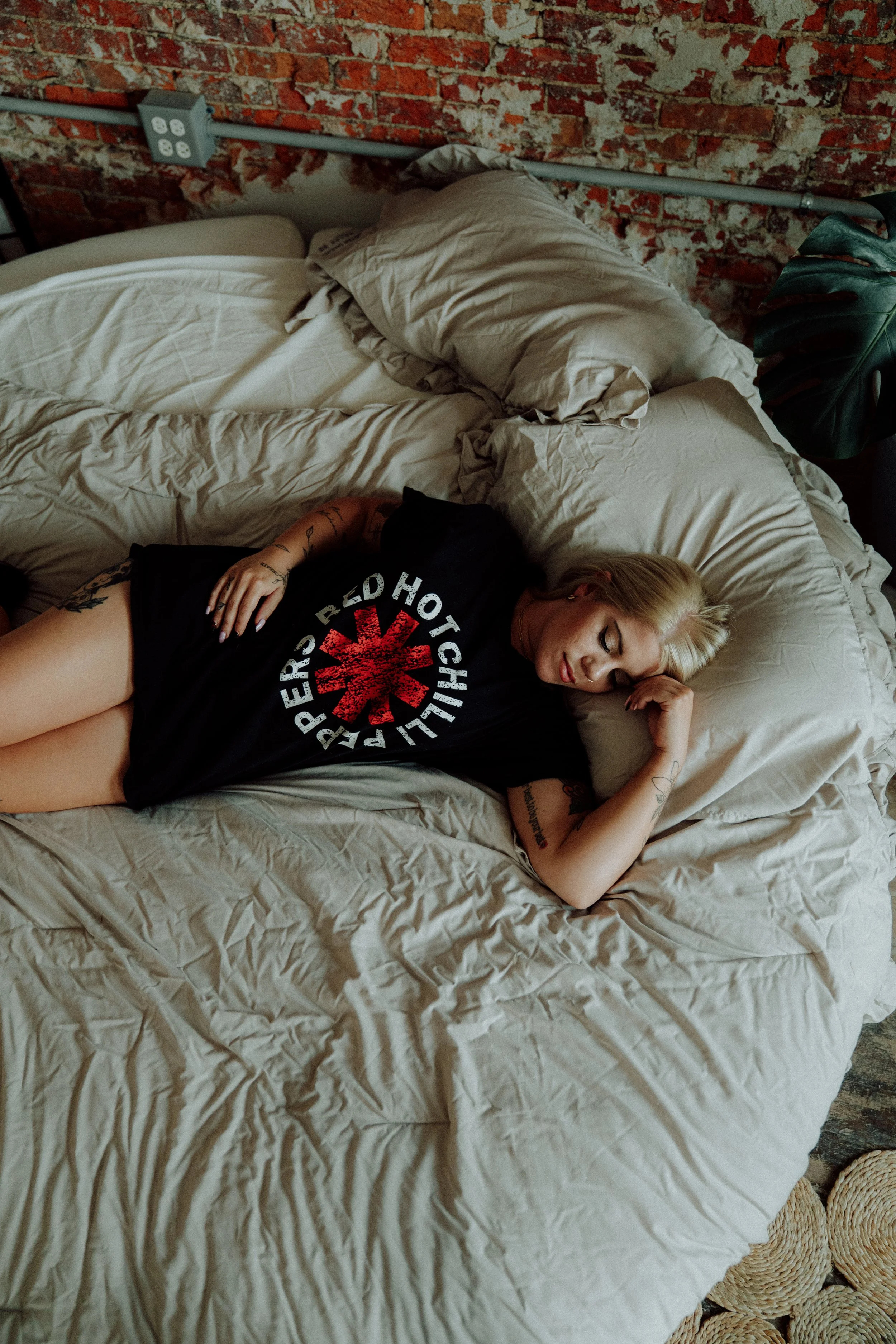 A woman with blonde hair and tattoos lying on her side on a bed with beige sheets and pillows, in front of a brick wall. She is wearing a black band t-shirt and appears to be resting or sleeping.