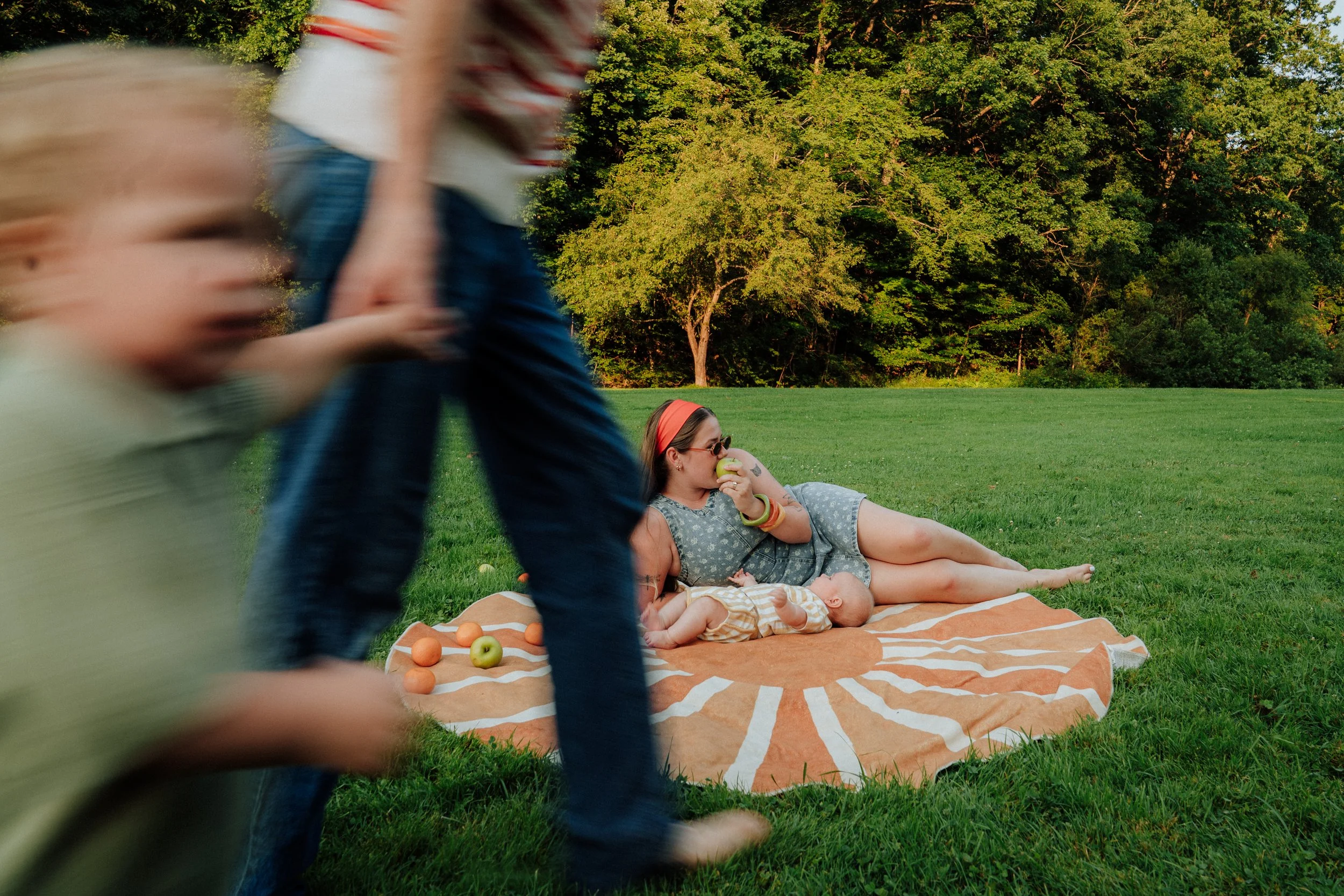 People walking past a woman lying on a blanket in a park with people playing and trees in the background.