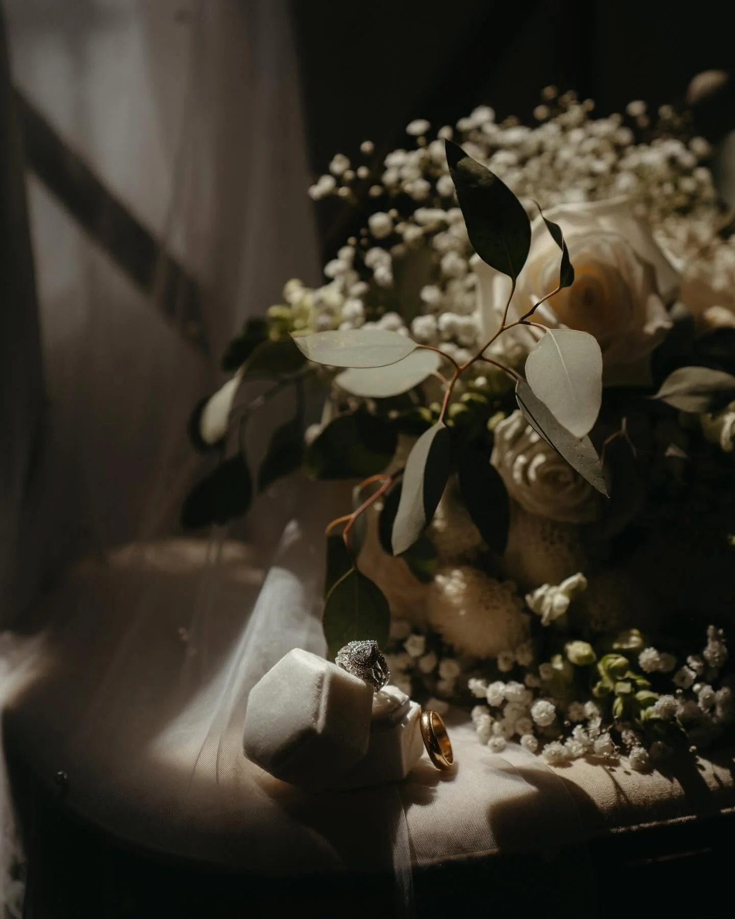 Close-up of a bouquet with white roses and green leaves, decorative rings and jewelry on a beige fabric surface with soft lighting.