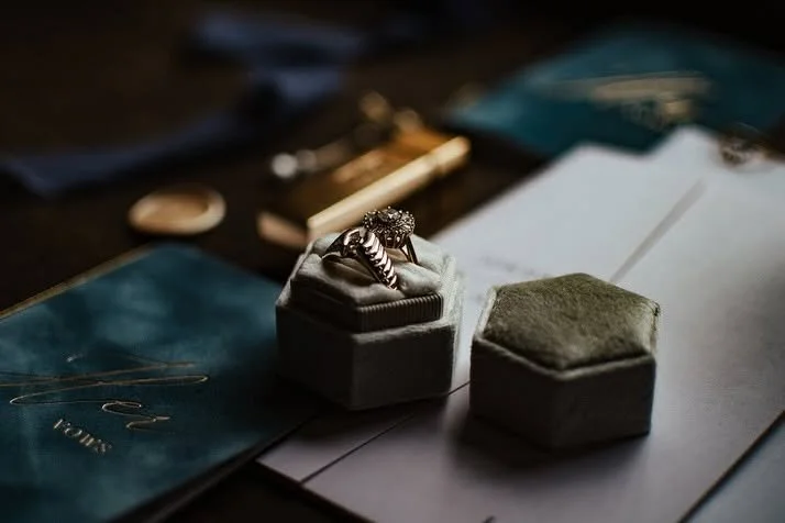 Close-up of a jewelry ring in a small gray box, with a green jewelry box beside it, on an open notebook with scattered documents and pen in the background.