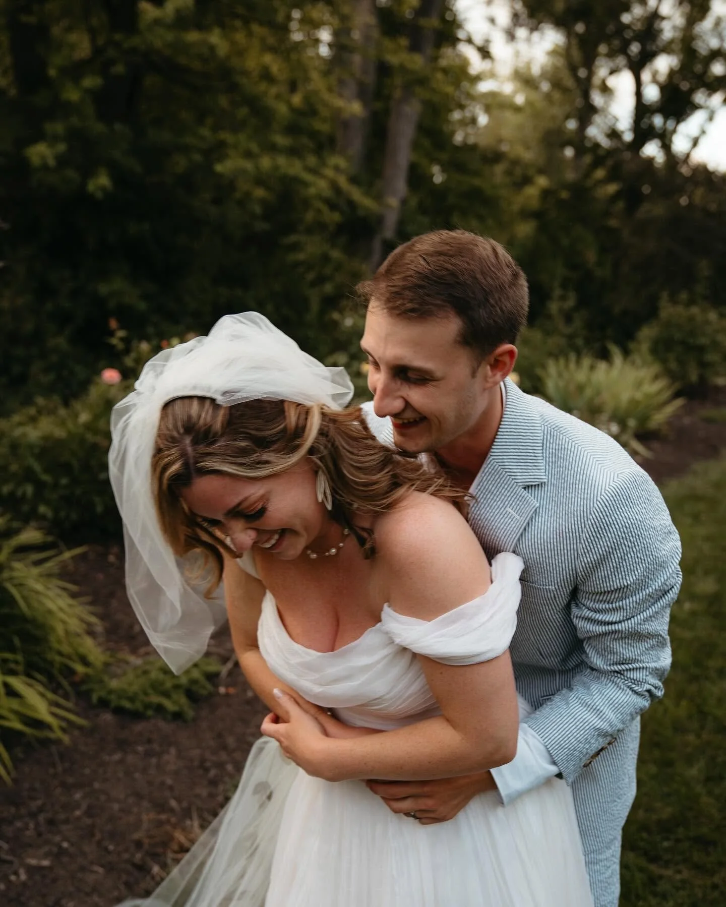 A bride and groom sharing a joyful moment outdoors, with the groom lifting the bride who is smiling and laughing, surrounded by trees and greenery.
