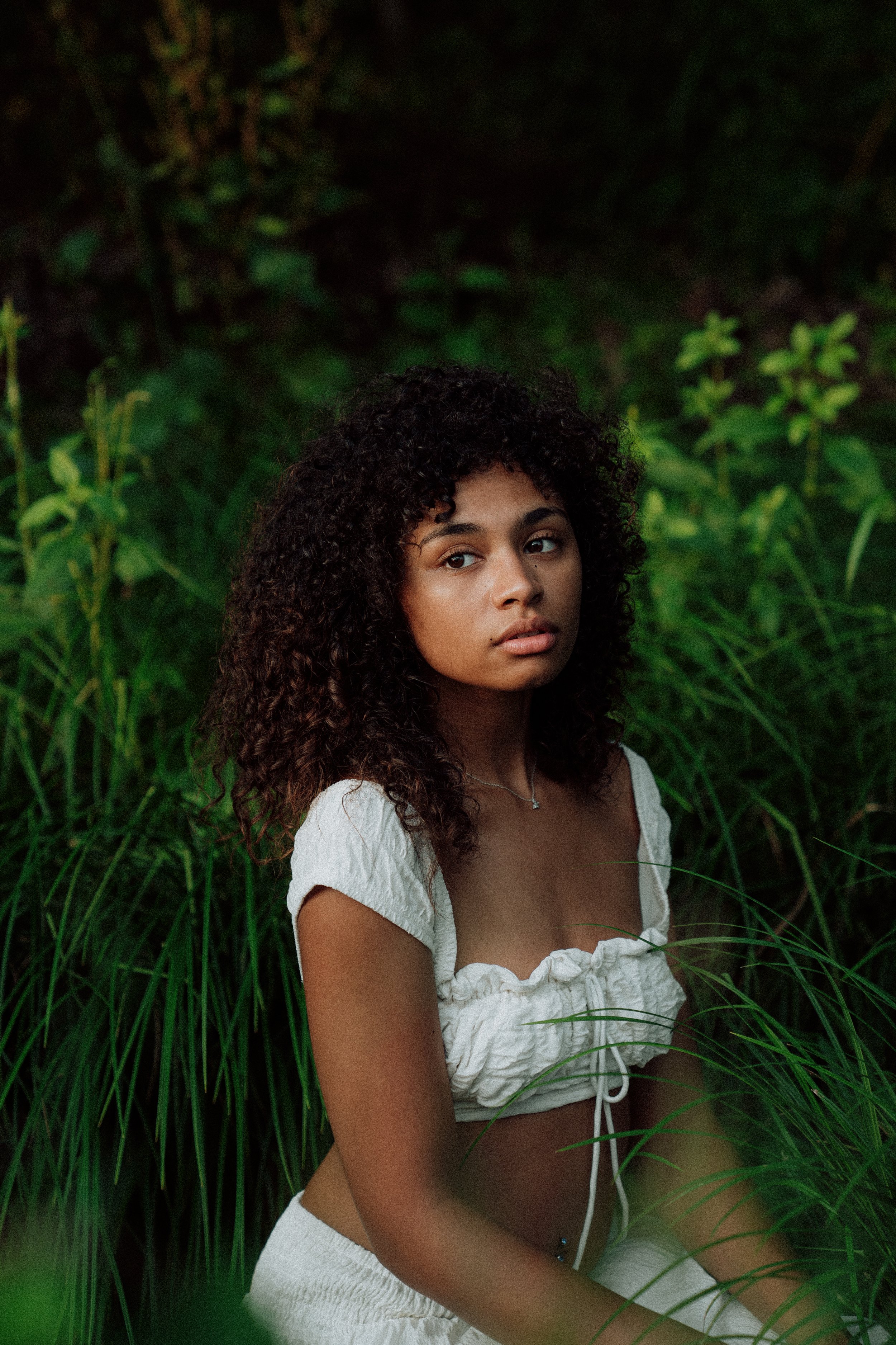 A young woman with curly hair, wearing a white cropped top and white shorts, sitting in a lush green outdoor setting surrounded by tall grass and plants.