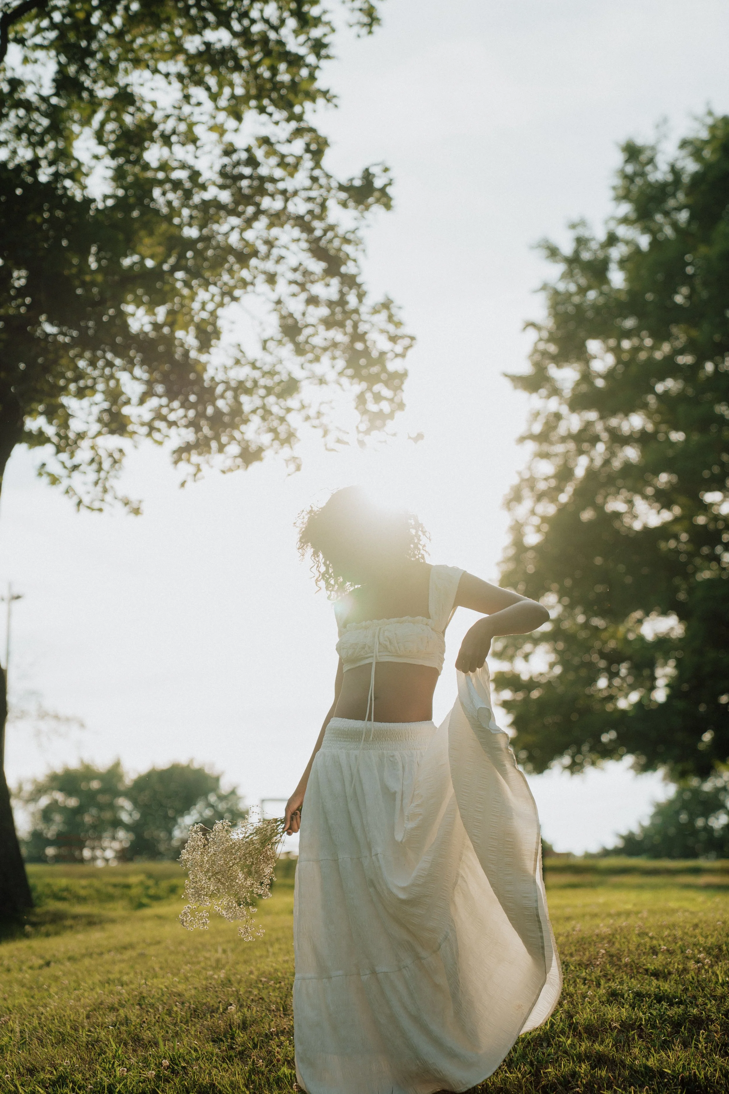 A woman in a white dress holding a bouquet of small white flowers standing outdoors in a grassy area with trees in the background, sunlight filtering through the trees.