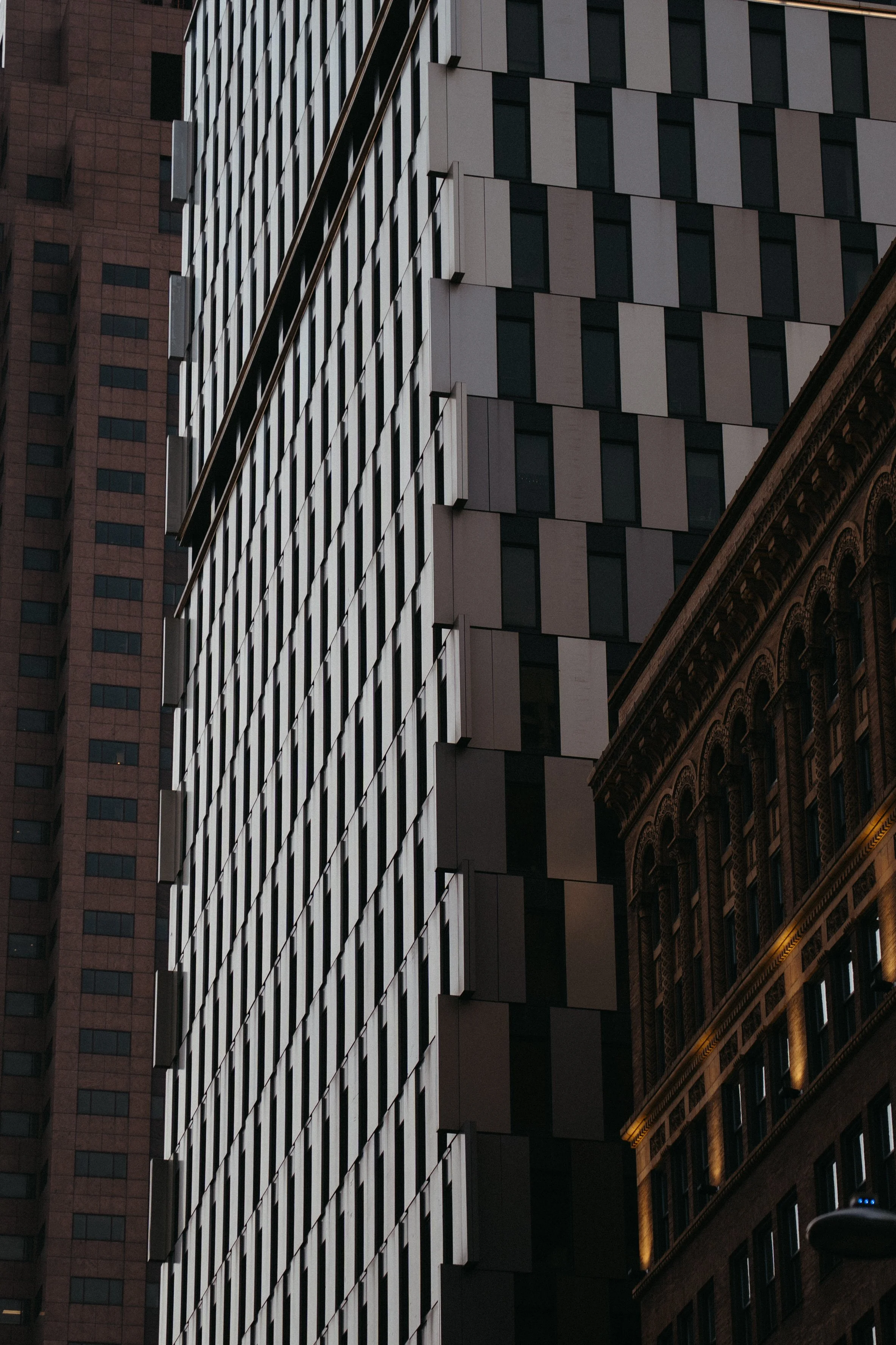 Image of a tall modern building with a facade made of rectangular panels and windows, with a portion of an older, ornate building visible at the bottom right.