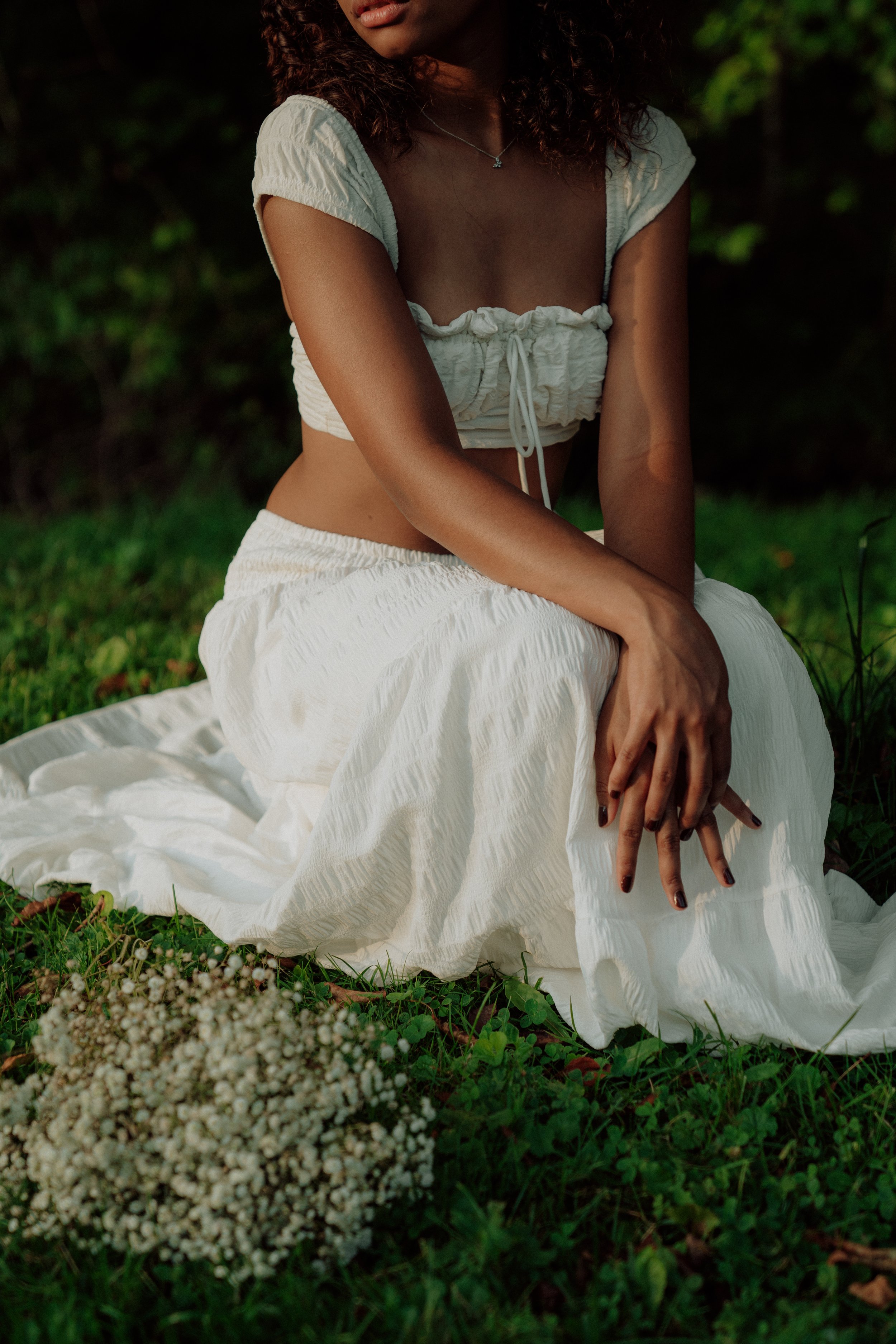 A woman sitting on the grass outdoors, wearing a white textured crop top with puffed sleeves and a long white skirt. She has curly hair and wears a silver necklace. In the foreground, there are small white flowers among green grass.