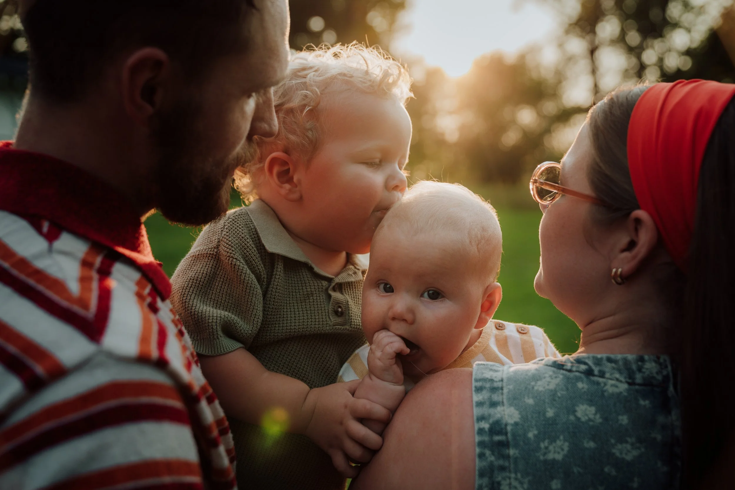 A family of five, including two women, a man, a young boy, and a baby, are close together outdoors during sunset, with trees and sunlight in the background, sharing an intimate moment.