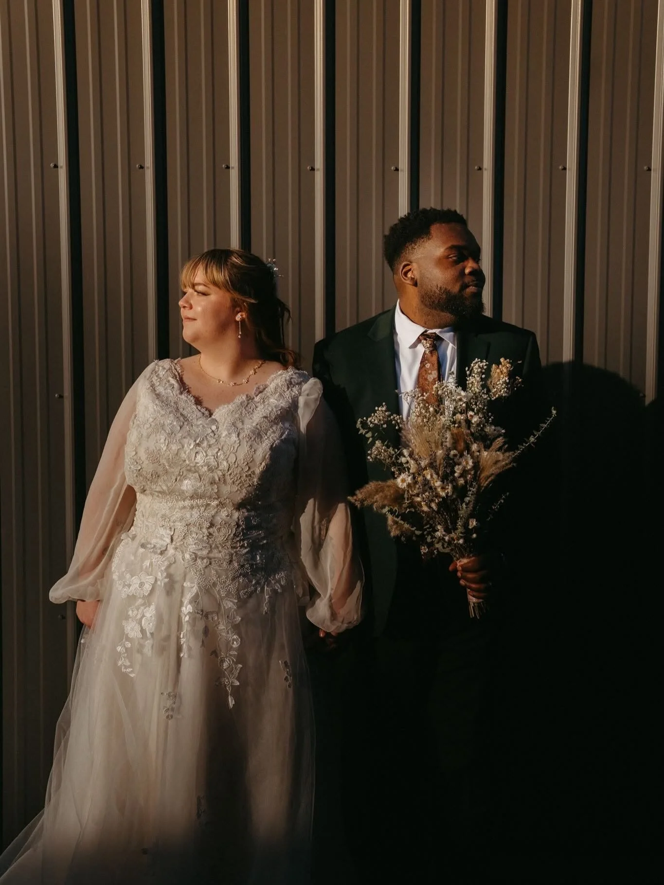 A bride and groom standing side by side against a metal wall, with the bride in a white lace wedding dress and the groom in a dark suit holding a bouquet of flowers.
