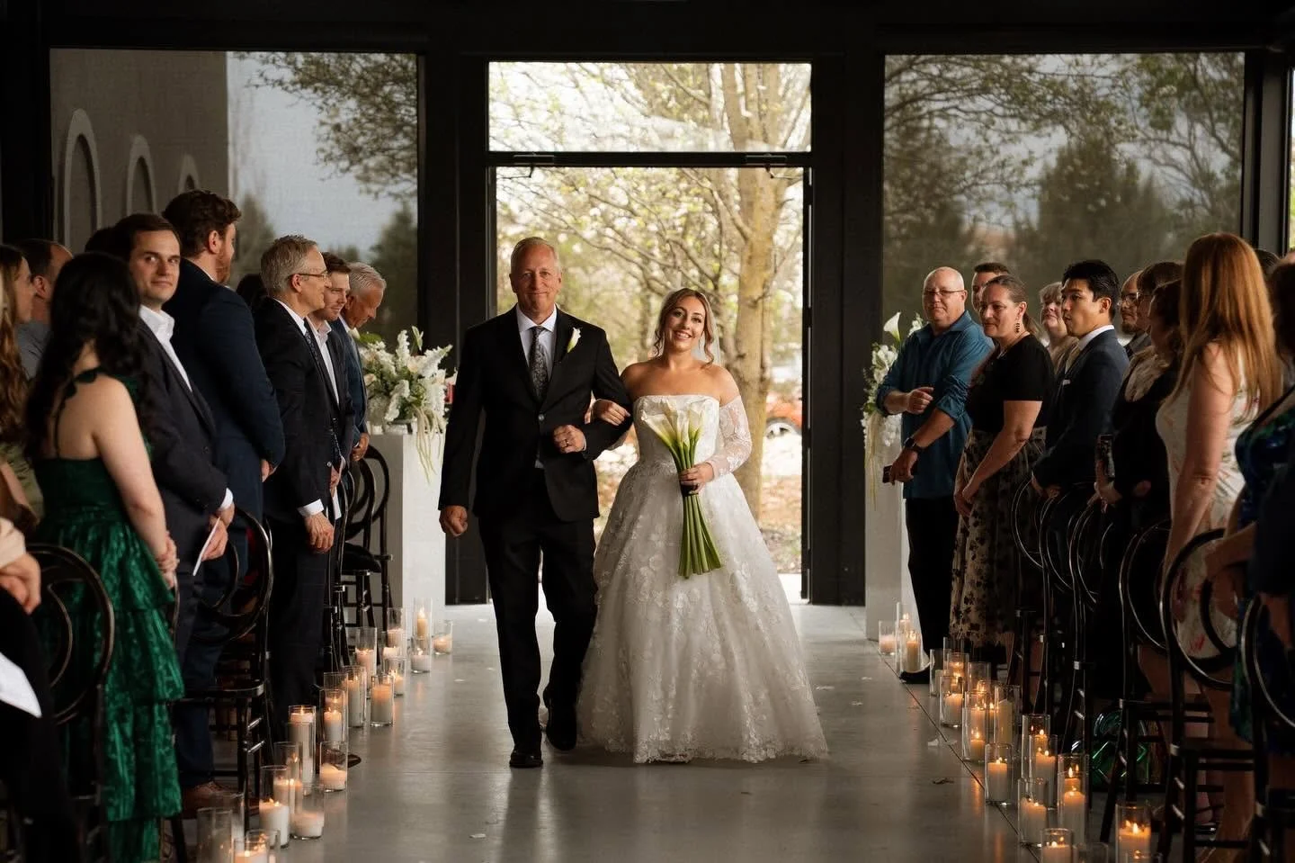 Bride walking down the aisle with her father during a wedding ceremony in a modern indoor venue, with guests on either side and candles lining the aisle.