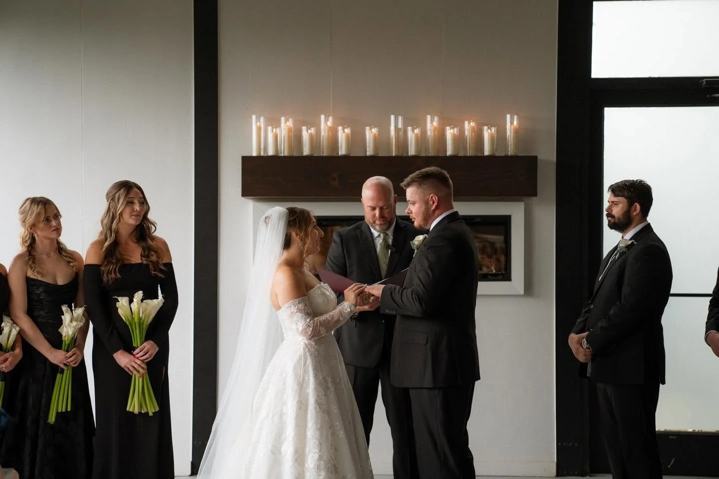 Bride and groom exchanging wedding vows during ceremony, with bridesmaids holding calla lilies standing nearby, inside modern indoor venue with candles on mantel.
