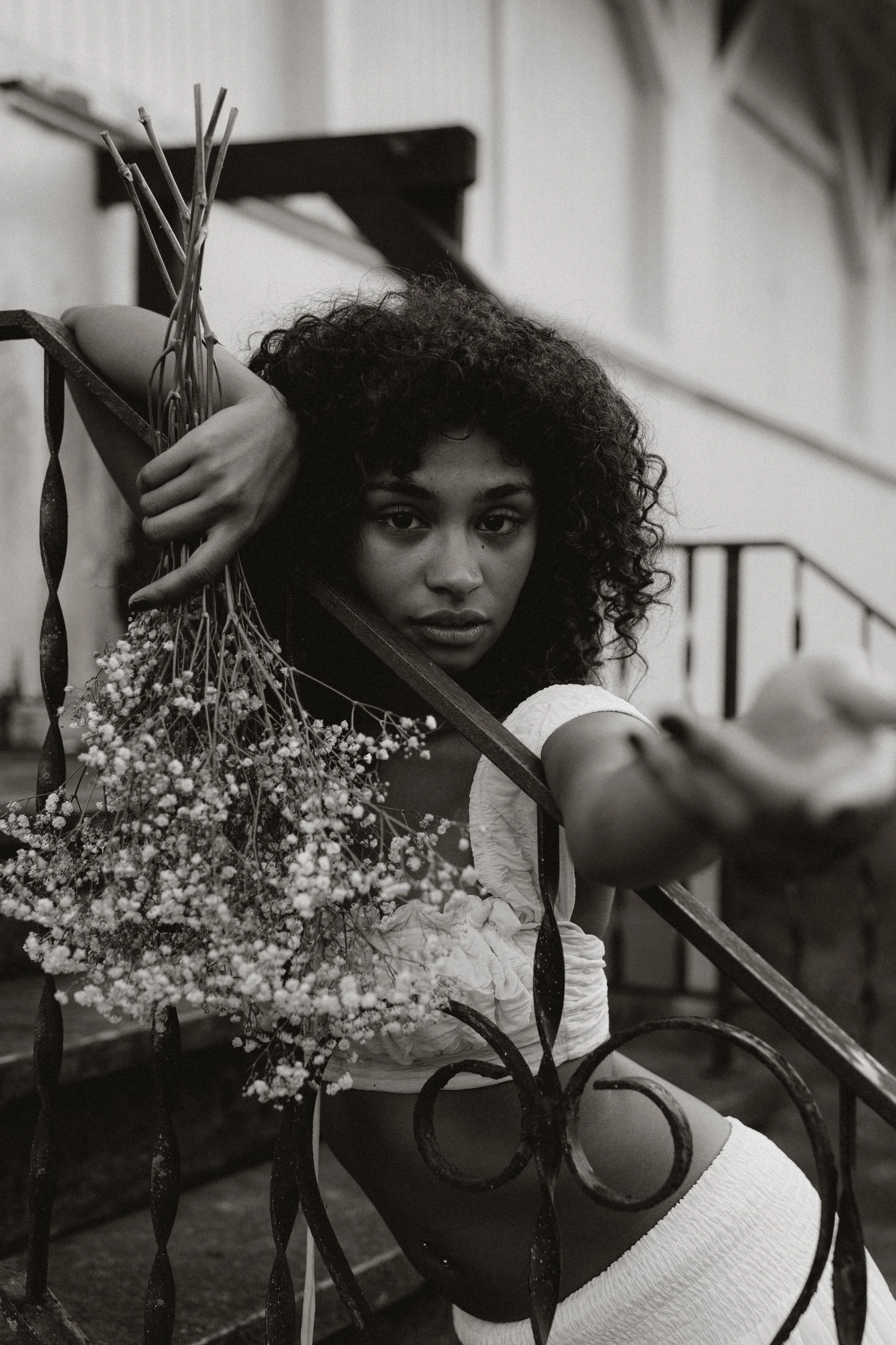 A woman with curly hair leaning on a decorative iron railing while holding a bouquet of flowers. She is looking directly at the camera with a calm expression.