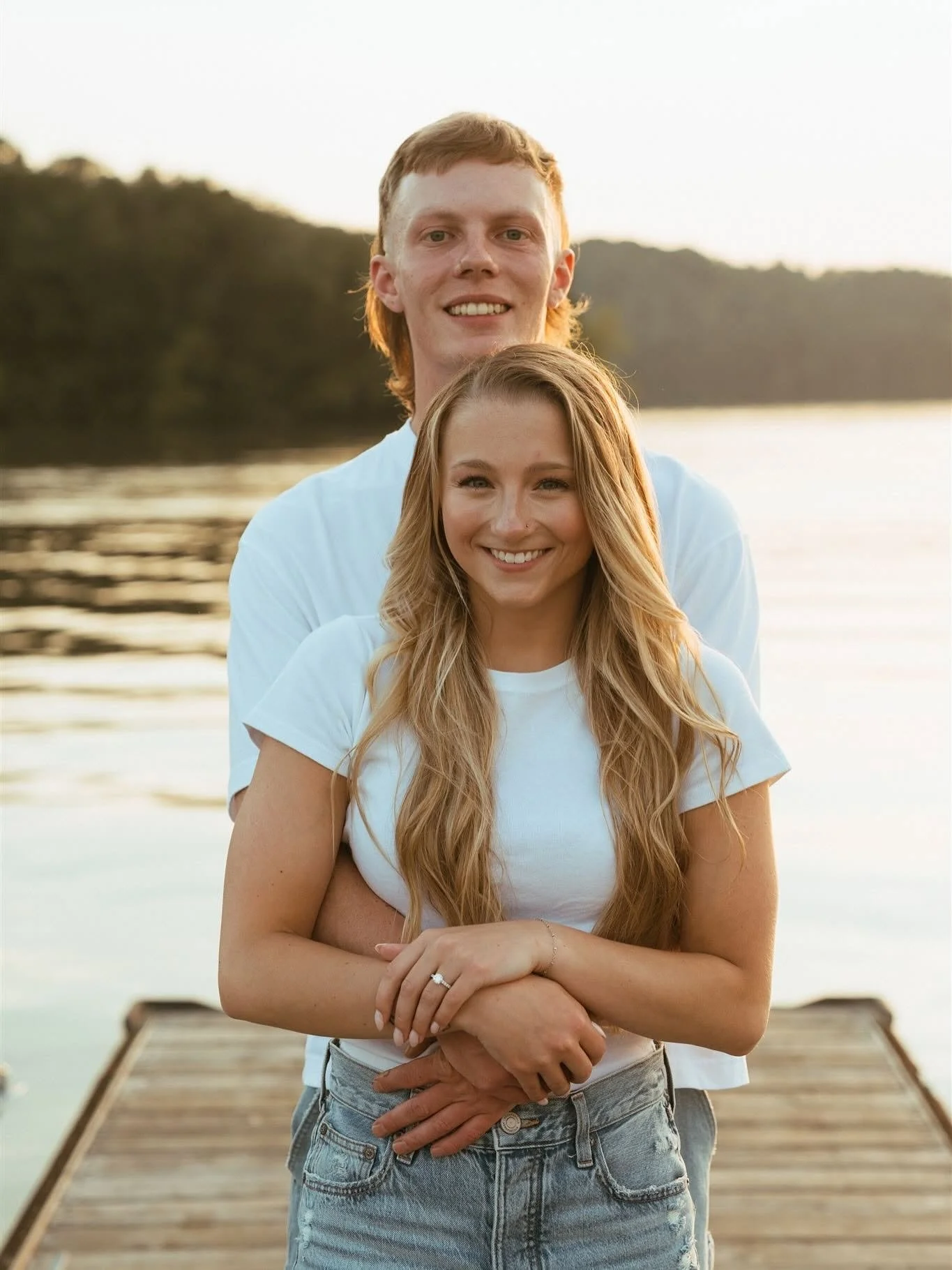 A young couple standing on a wooden dock by a lake at sunset, smiling at the camera. The man is behind, wearing a white shirt, and the woman is in front, wearing a white t-shirt and jeans, with long blond hair, holding her hands together.