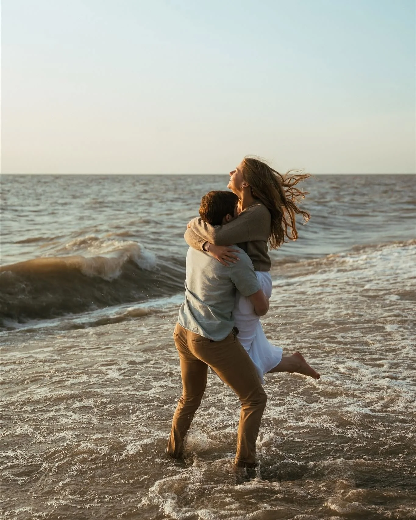 A man and woman embracing on the beach as the man lifts the woman off the ground. The woman has long hair and is wearing a gray sweater and white skirt, smiling with her eyes closed. The man is wearing a light-colored shirt and brown pants, holding t