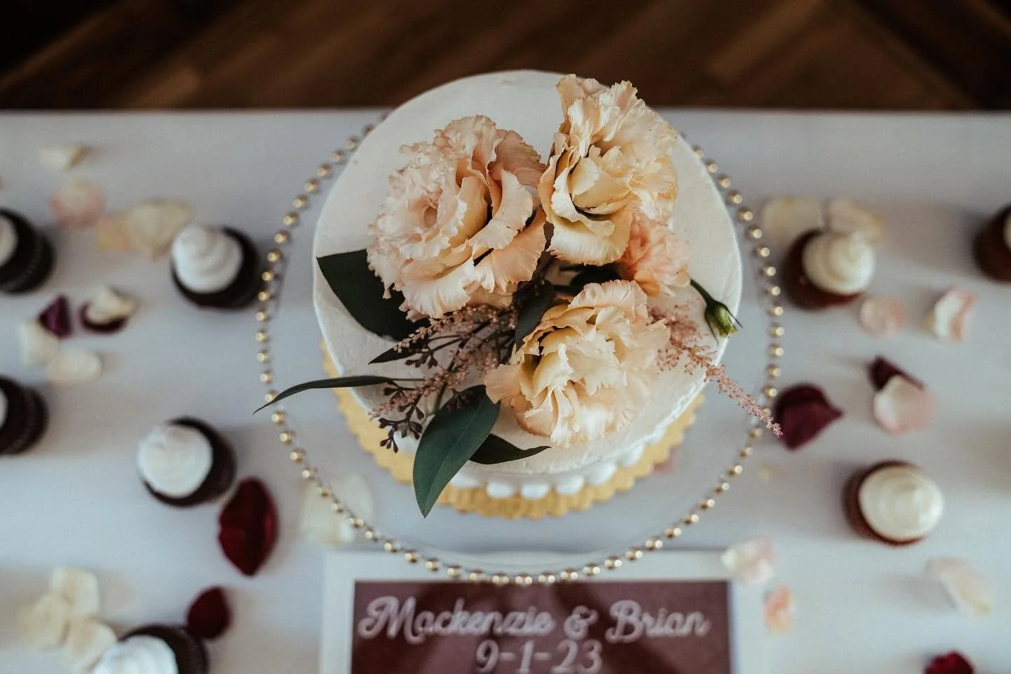 Decorated wedding cake topped with peach-colored flowers and greenery, surrounded by cupcakes and flower petals on a white table, with a sign reading "Mackenzie & Brian 9-1-23".