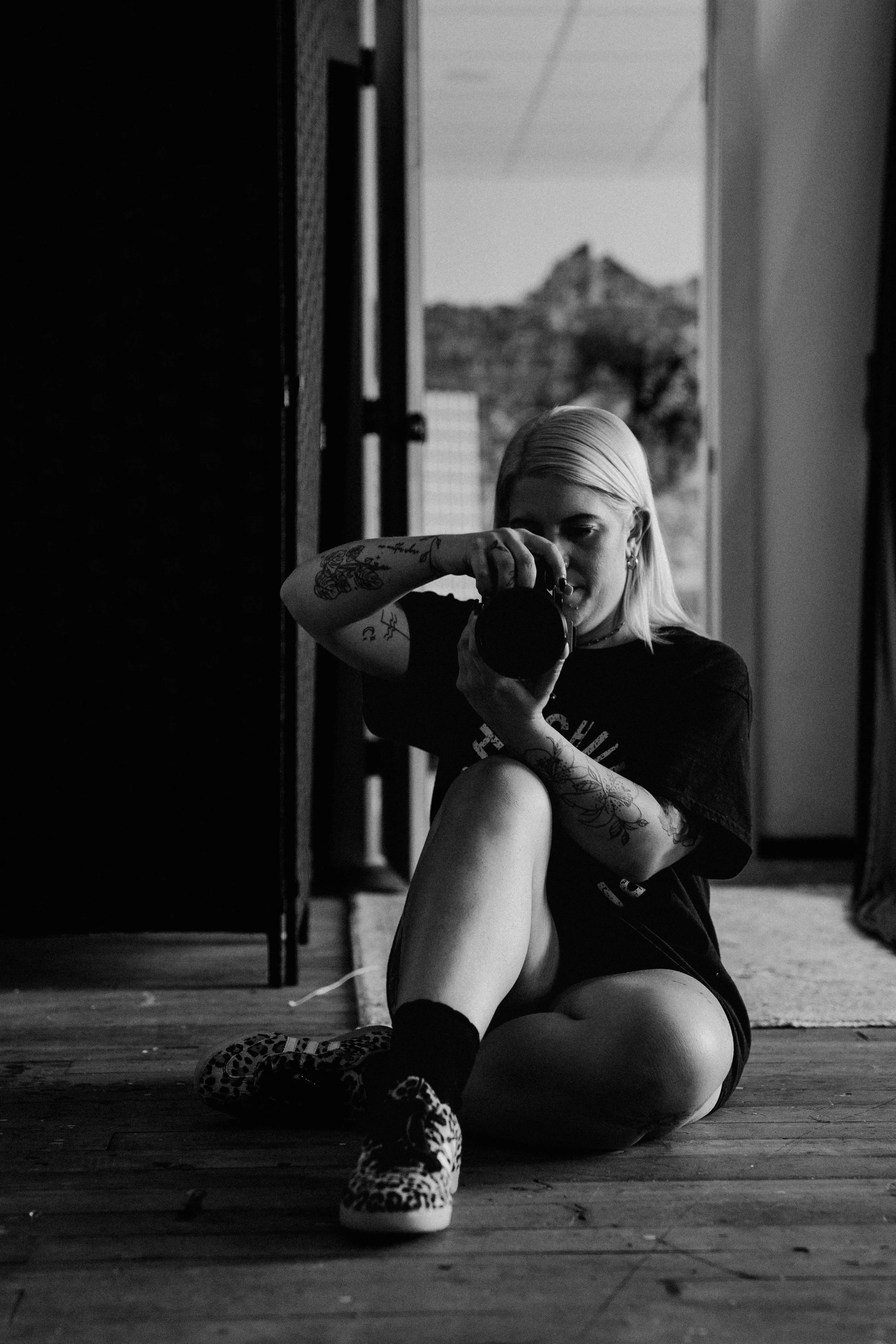 A woman with platinum blonde hair taking a selfie with a camera. She has tattoos on her arms and is sitting indoors on a hardwood floor, with a window and outdoor scenery visible in the background.
