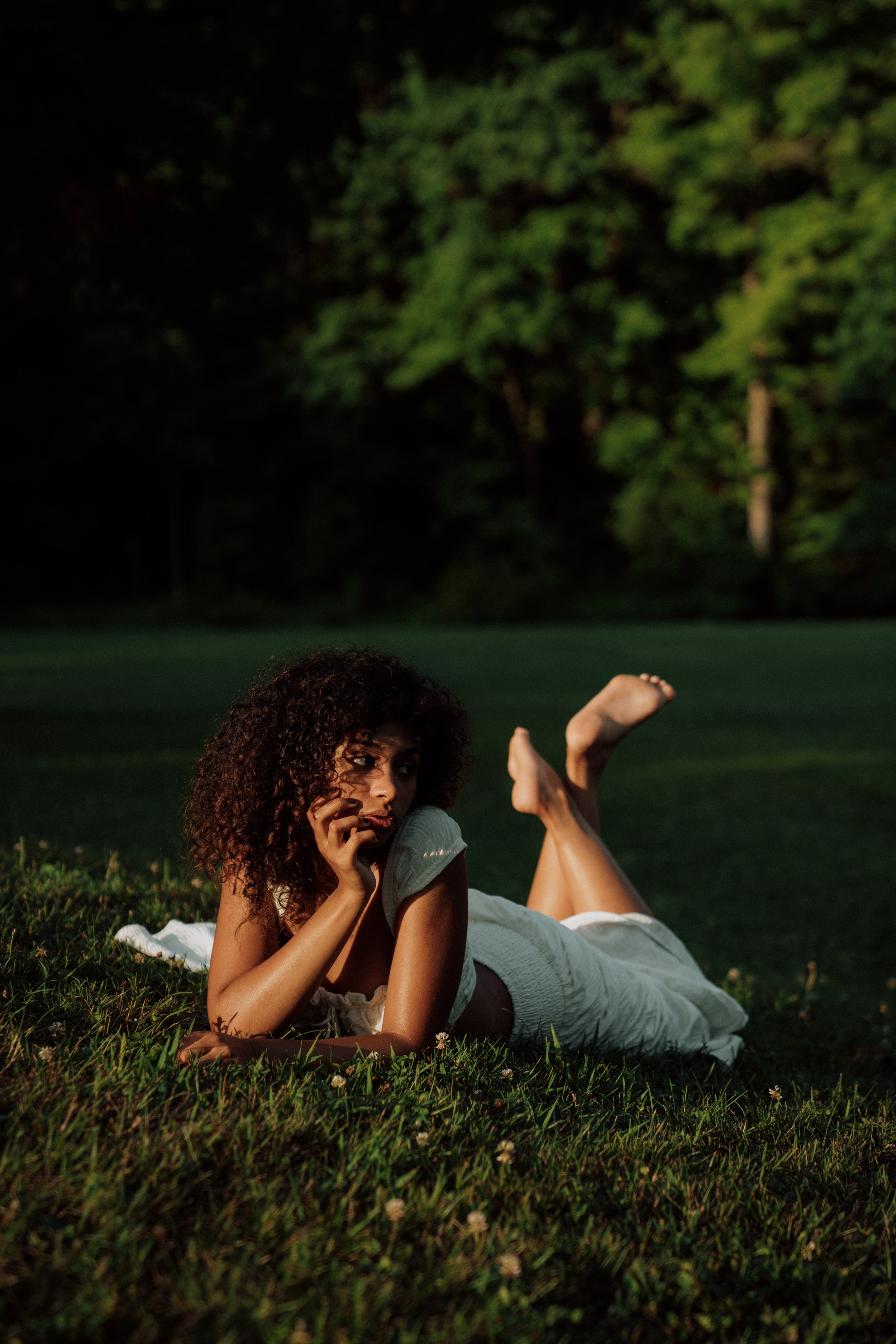 A woman lies on her side on a grassy field at night, with trees in the background, holding her chin with one hand and resting her cheek on the other, looking thoughtfully into the distance.
