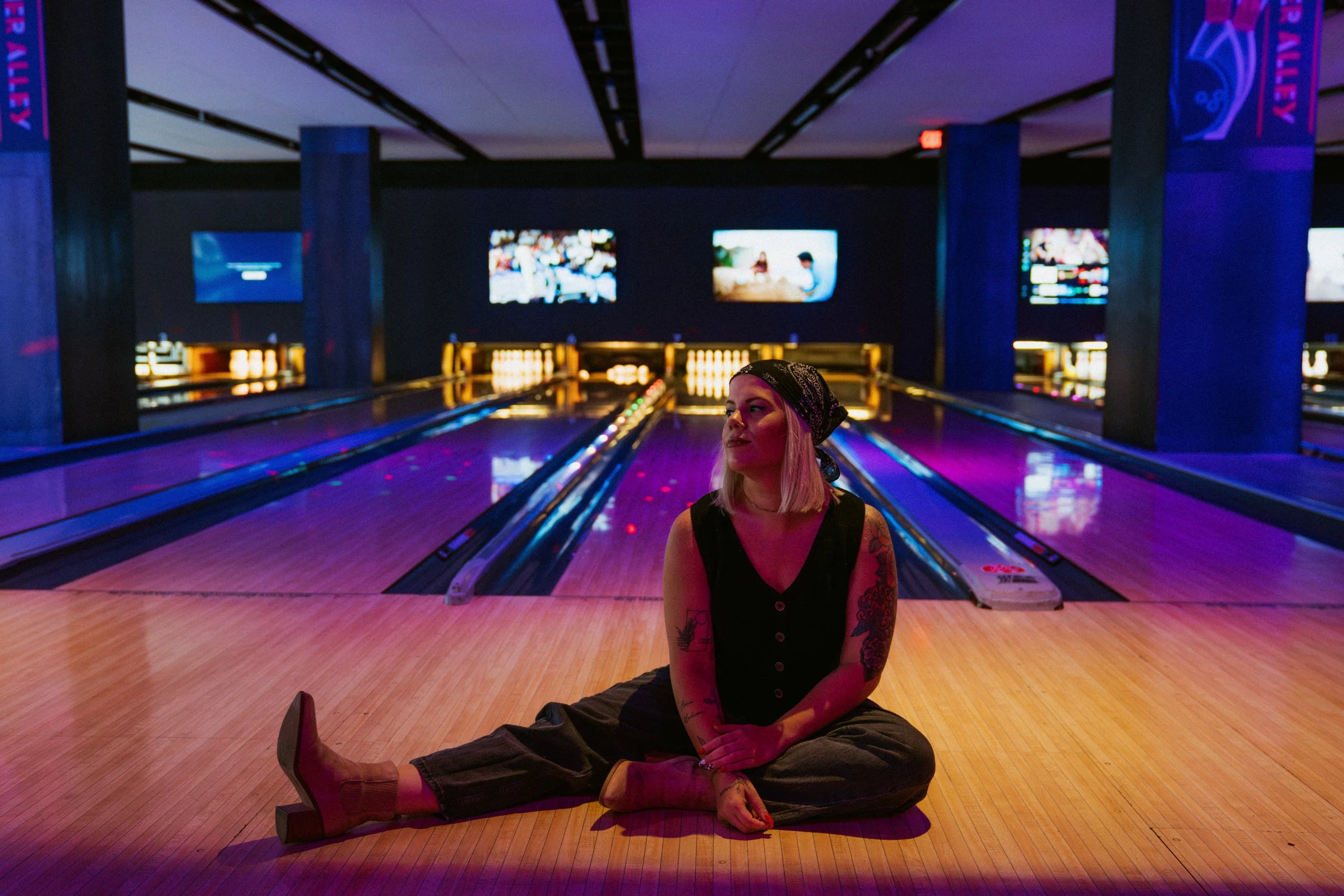 A woman with tattoos, sitting cross-legged on a bowtie-shaped mat in a dimly lit bowling alley with purple and pink neon lighting, wearing a bandana and casual clothing.