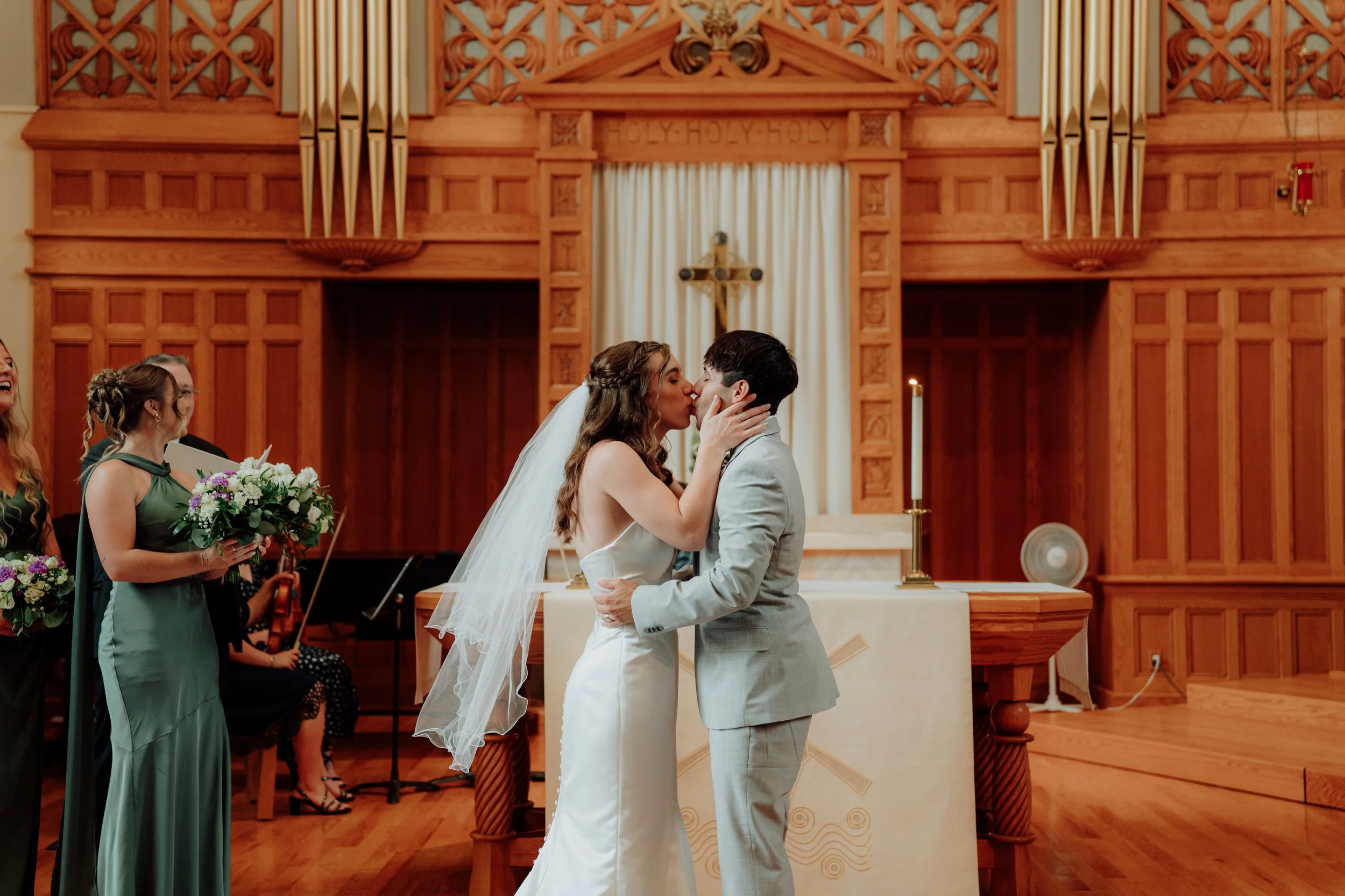 A bride and groom kiss during their wedding ceremony in a church, surrounded by bridesmaids holding bouquets.