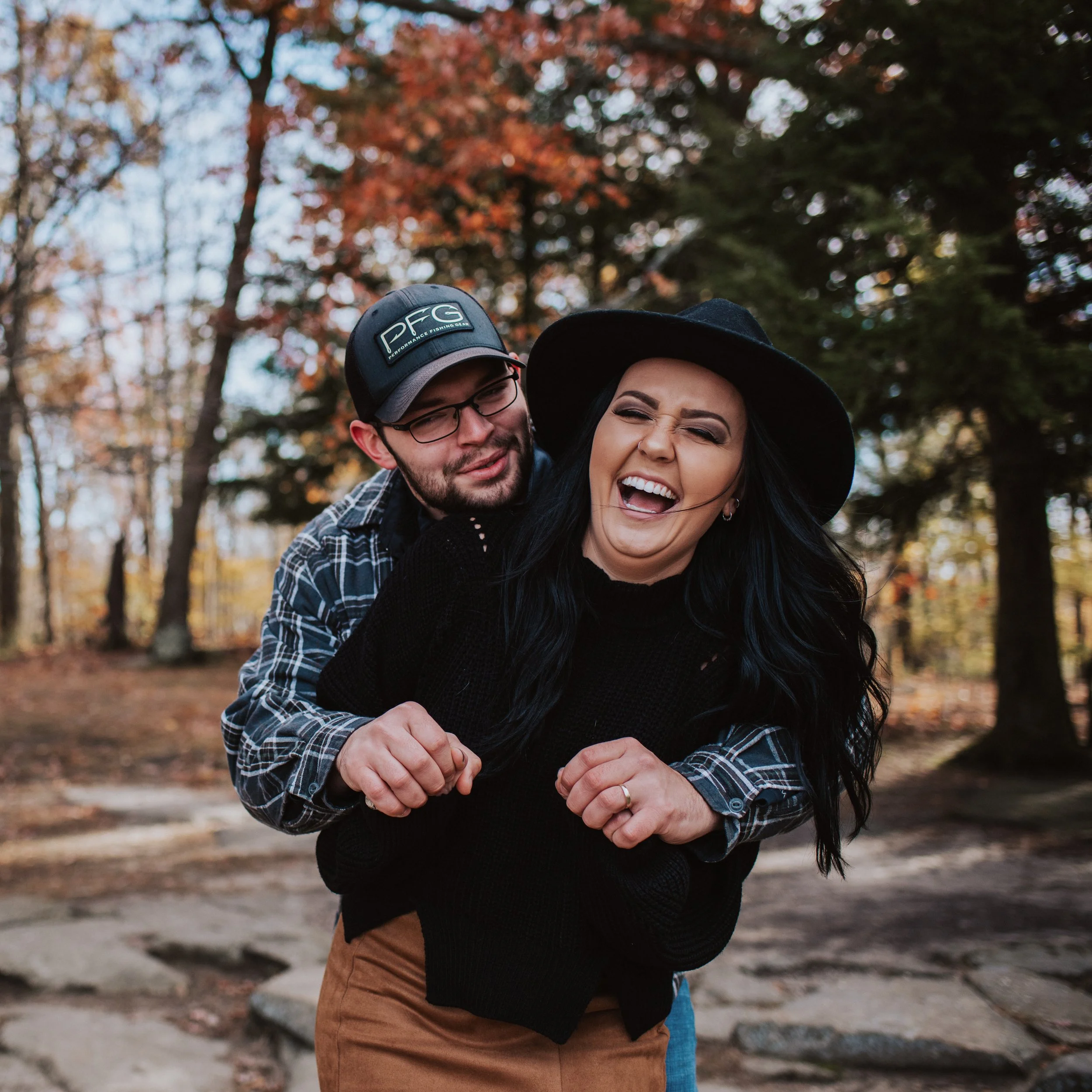 Close-up of two people, a man and a woman, smiling and hugging outdoors during autumn with trees with red and yellow leaves in the background.