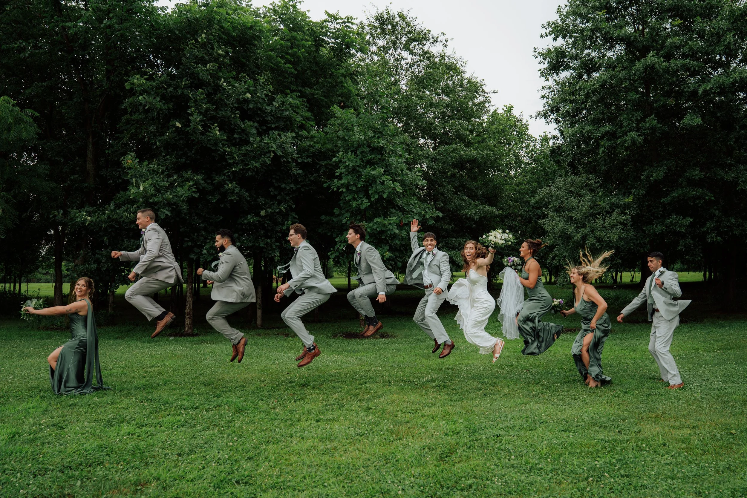 Wedding party jumping in a line outdoors surrounded by trees, with women in formal dresses and men in suits.