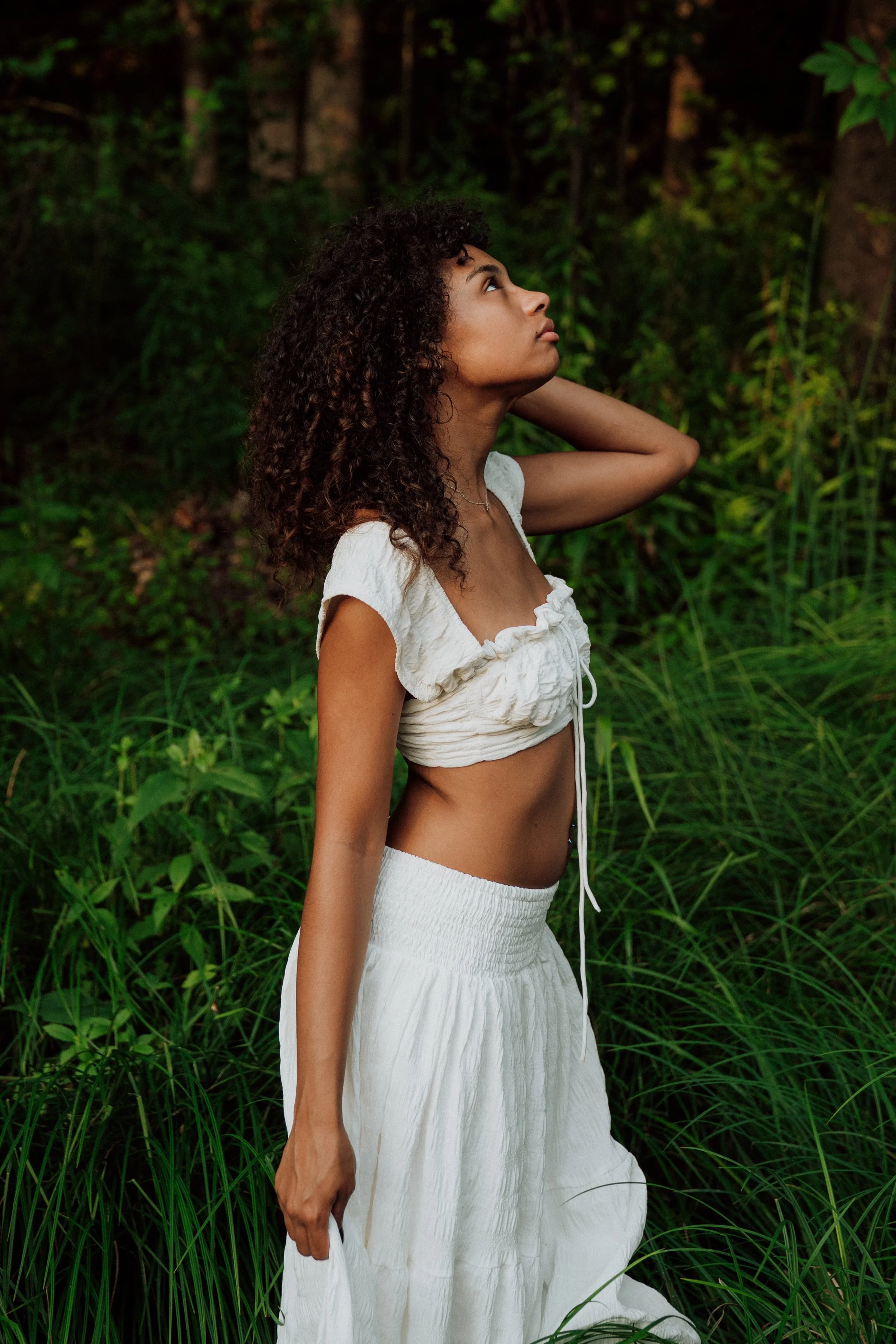 A woman with curly hair standing in a green, forested area. She is wearing a white, ruffled crop top and a long white skirt, looking upwards with one hand behind her neck.