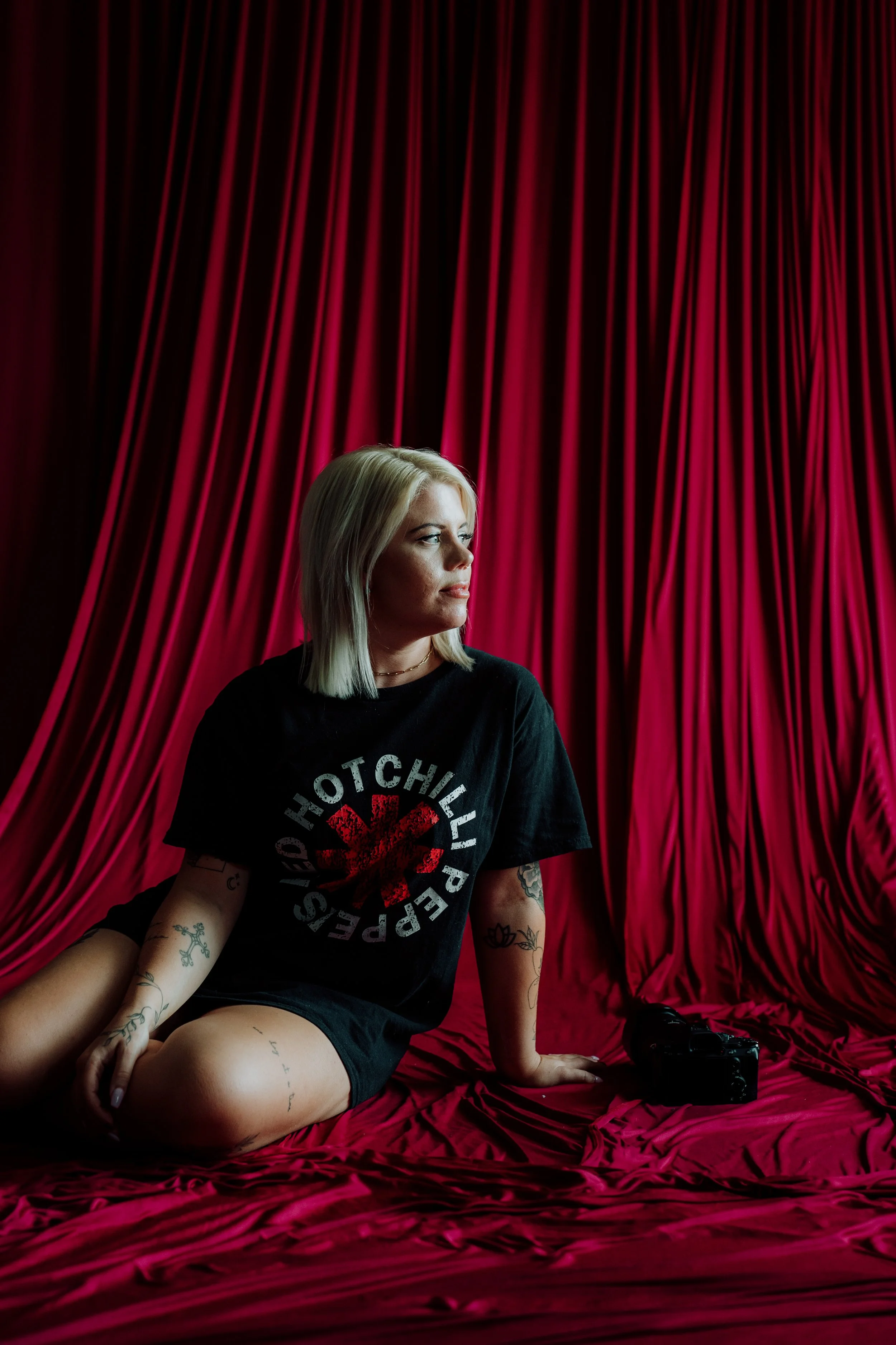 A woman with blonde hair sitting on a red silky fabric backdrop, wearing a Black Flag t-shirt and black shorts, with various tattoos on her arms and legs, holding a camera.