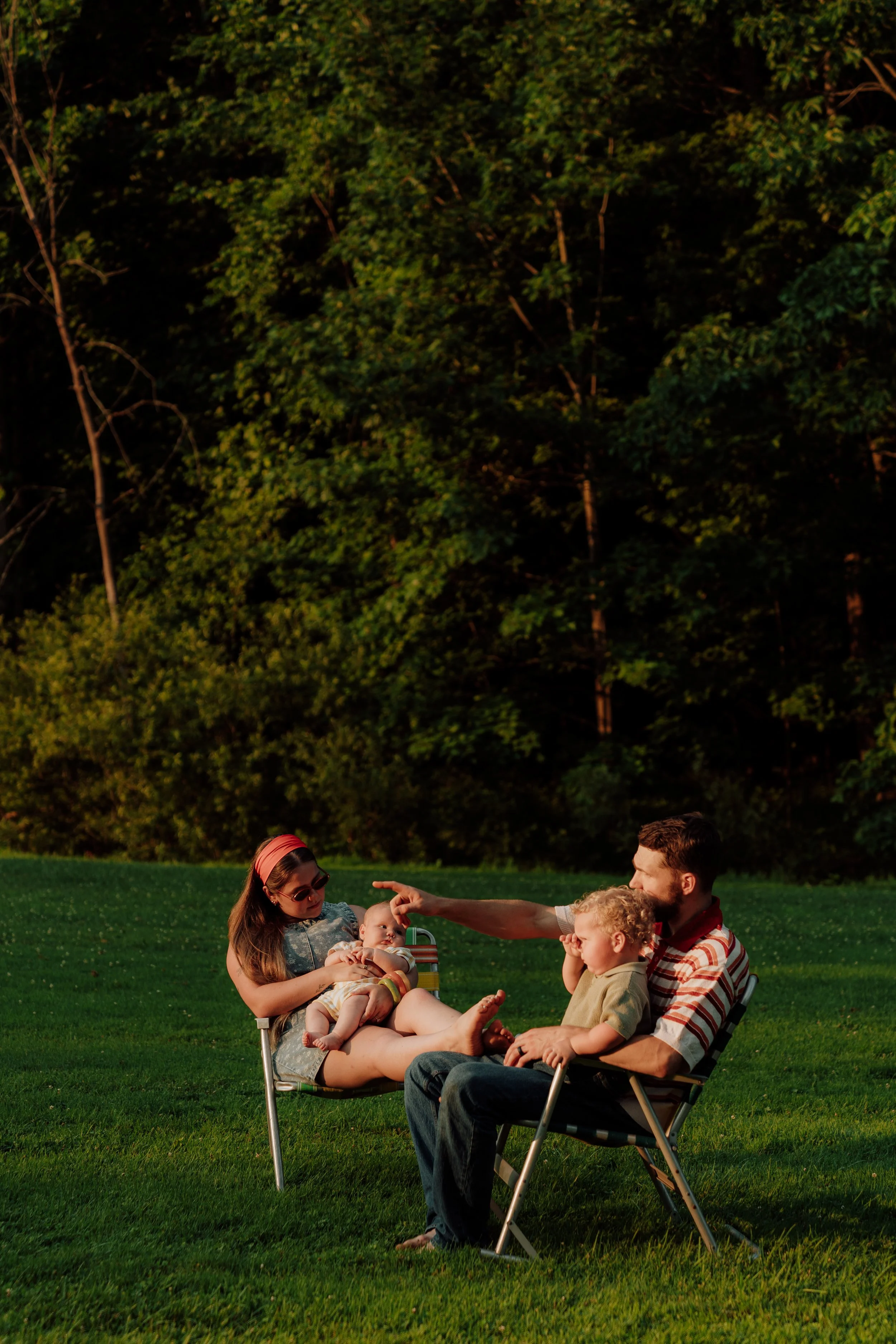 Family of five sitting on folding chairs on a grassy field, with trees in the background. The woman is holding a baby, and the man and two children are engaged in conversation, with sunlight casting a warm glow.
