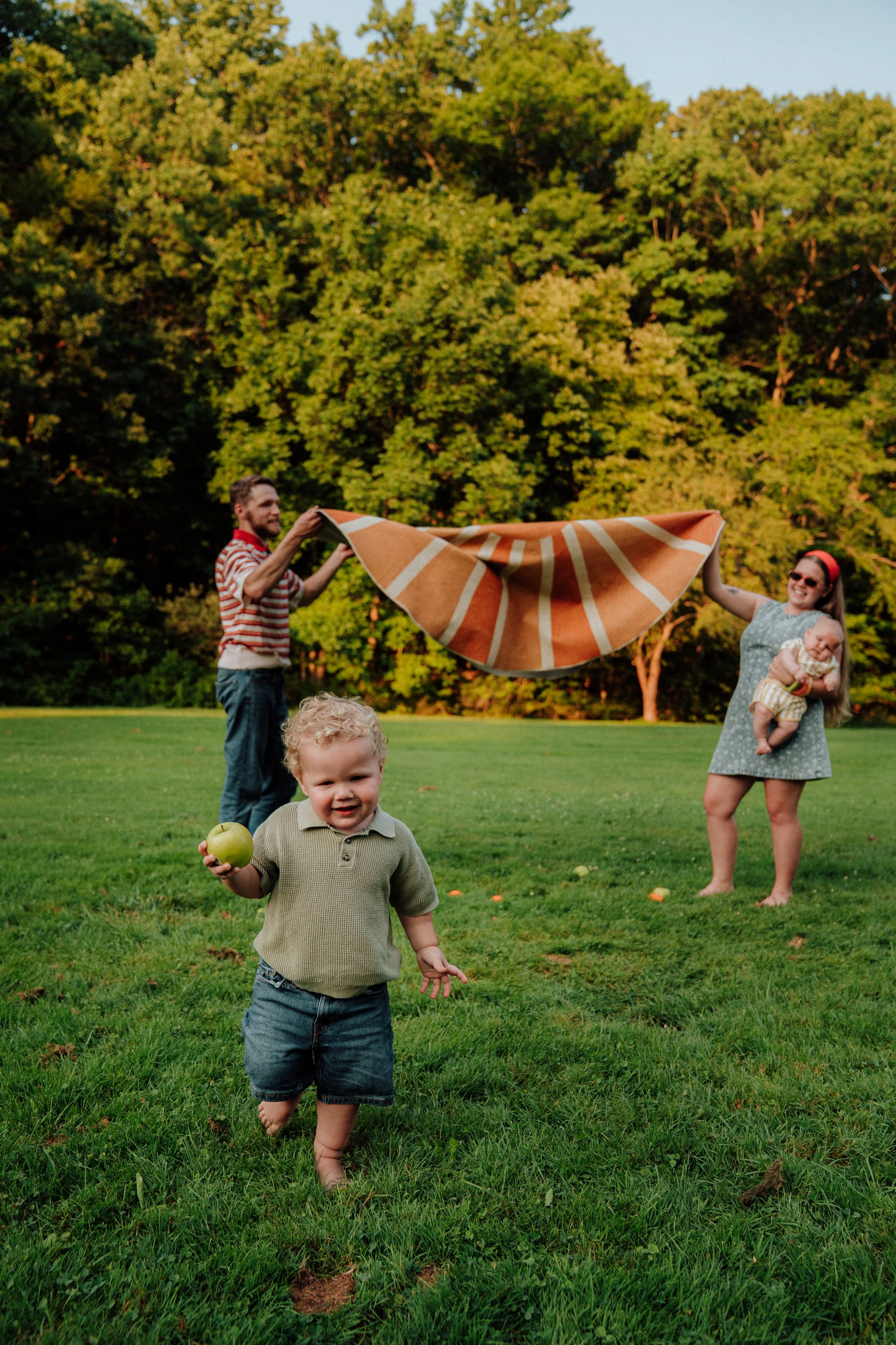 A young boy with blonde curly hair and a green polo shirt, holding a green apple, walking barefoot on a grassy field. In the background, a woman with sunglasses and a red headband holds a baby, and a man in a striped shirt is lifting a large orange a