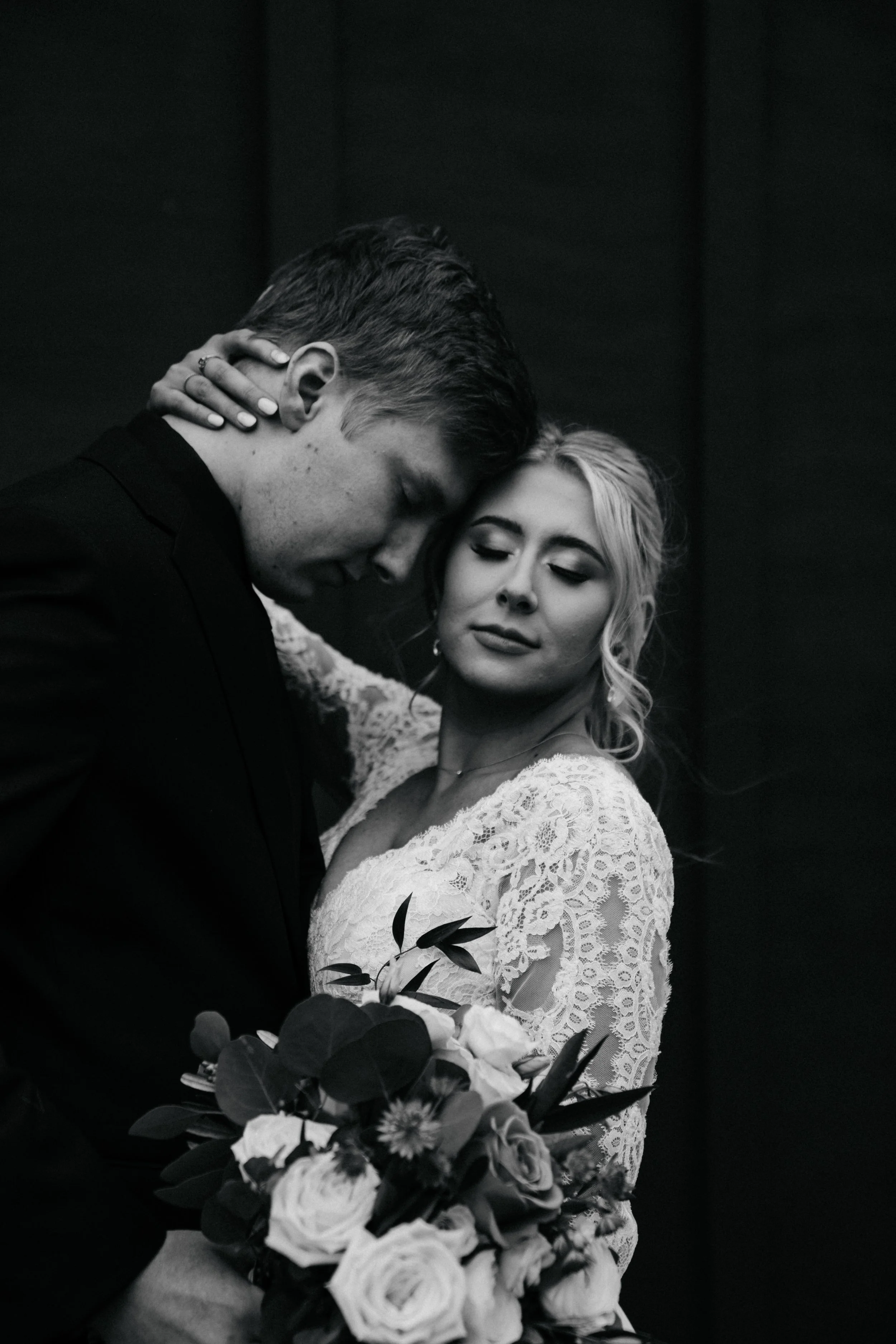 Black and white photo of a loving couple, with the man kissing the woman's forehead, she with closed eyes, holding a bouquet of flowers.